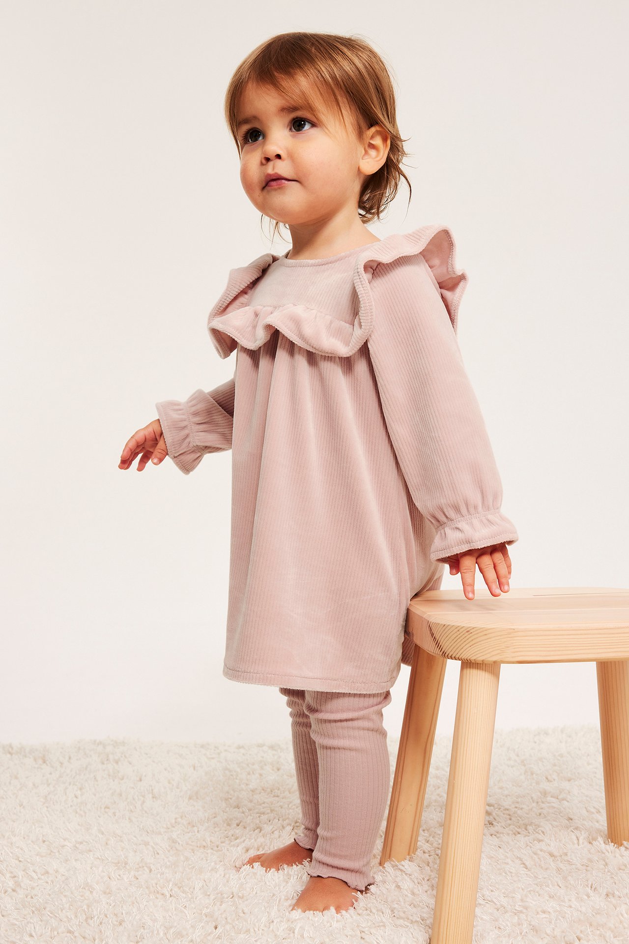 Forfra: baby i rosa ribbet velourkjole med volanger og matchende rosa ribbestrikket leggings, berører krakk.