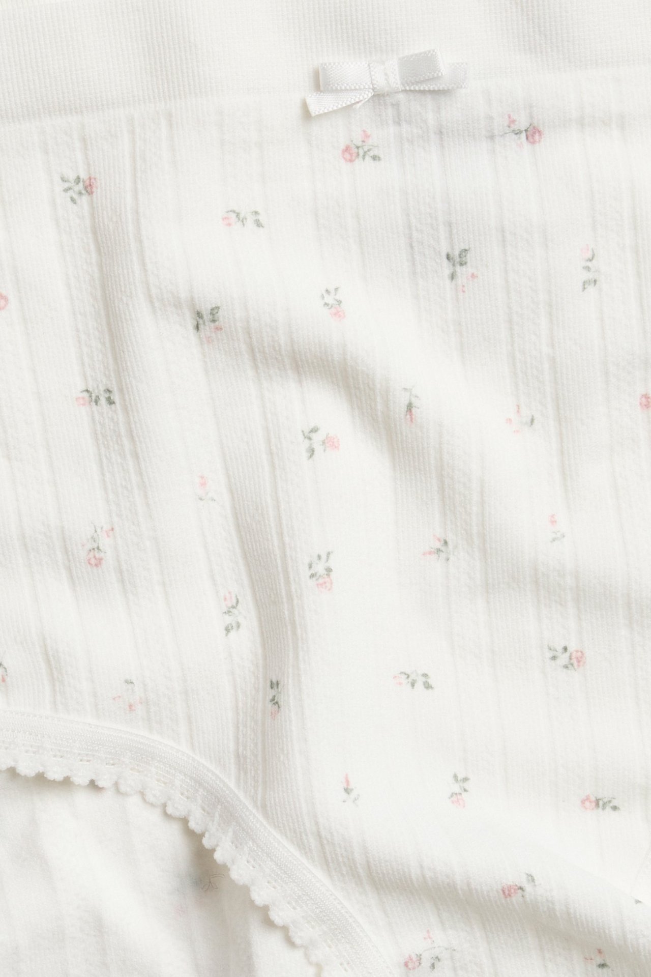 Zbliżenie: Białe, prążkowane, bezszwowe figi hipster z różowym kwiatowym wzorem, małą kokardką i obszyciem picot.