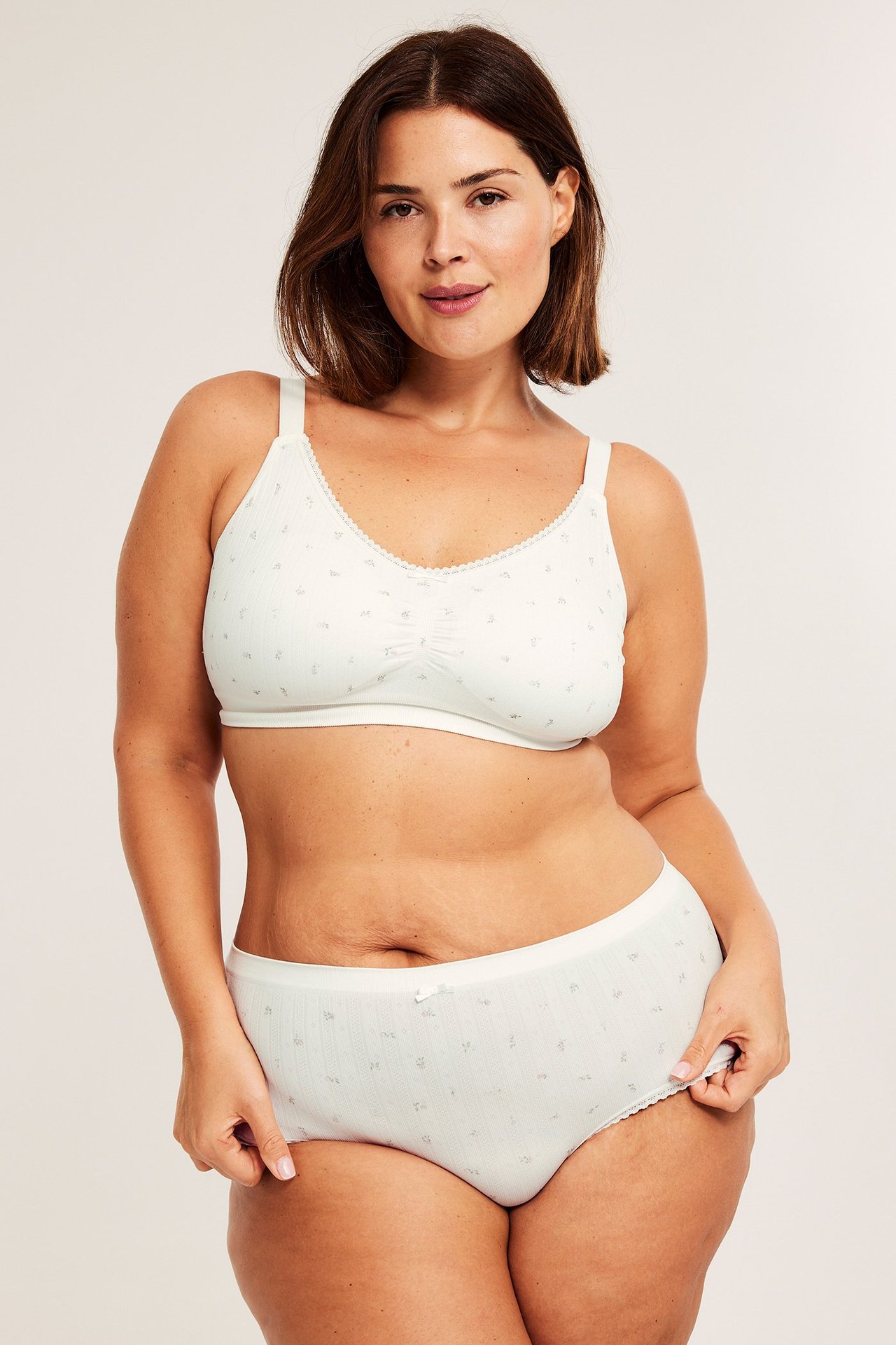 Frontutsikt av en kvinne i kremhvite blomstrete hipstertruser med sløyfe, og matchende BH.