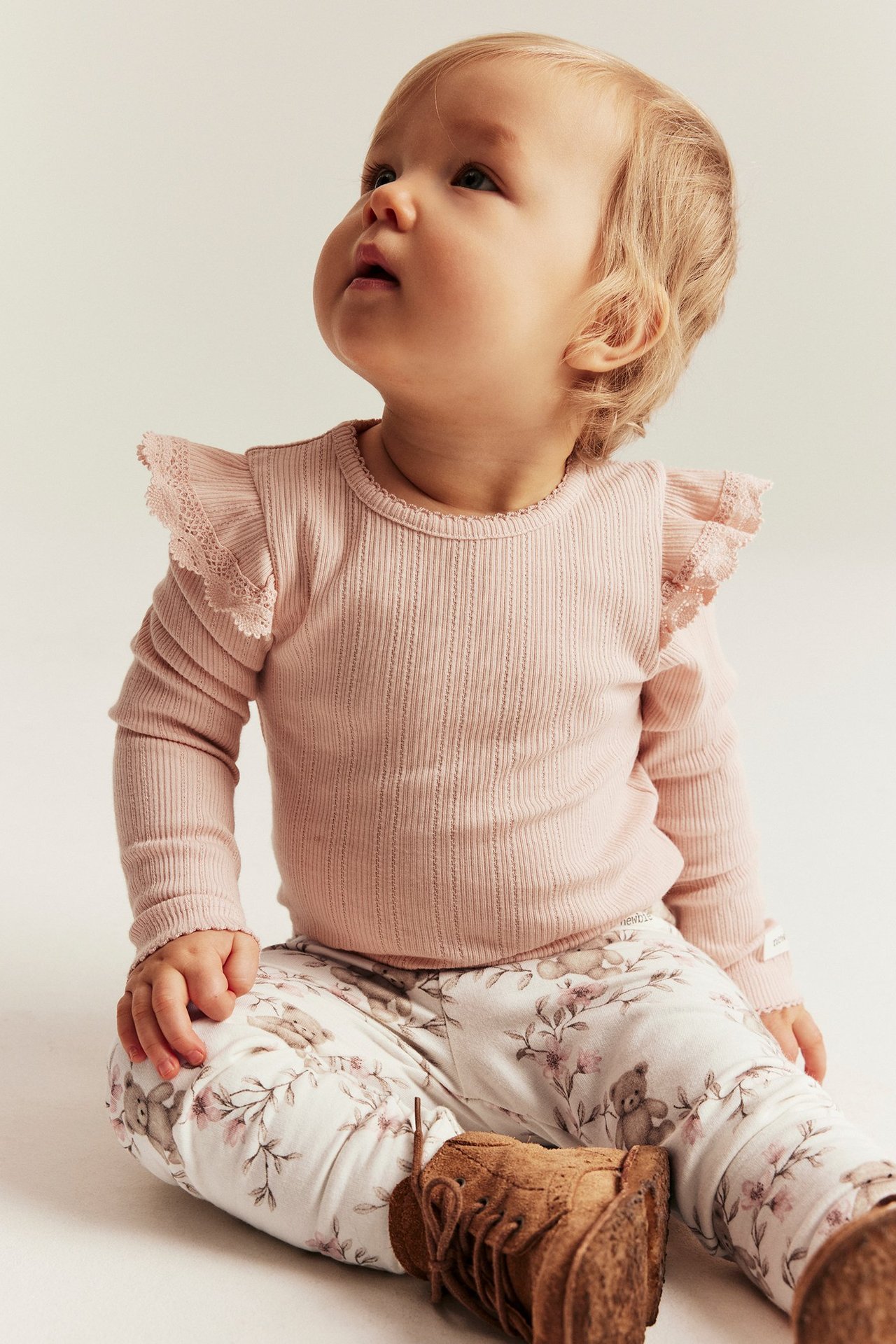 Frontvisning av baby i kremhvite blomstrete leggings med teddybjørner, rosa ribbet topp og brune frynsede støvler.