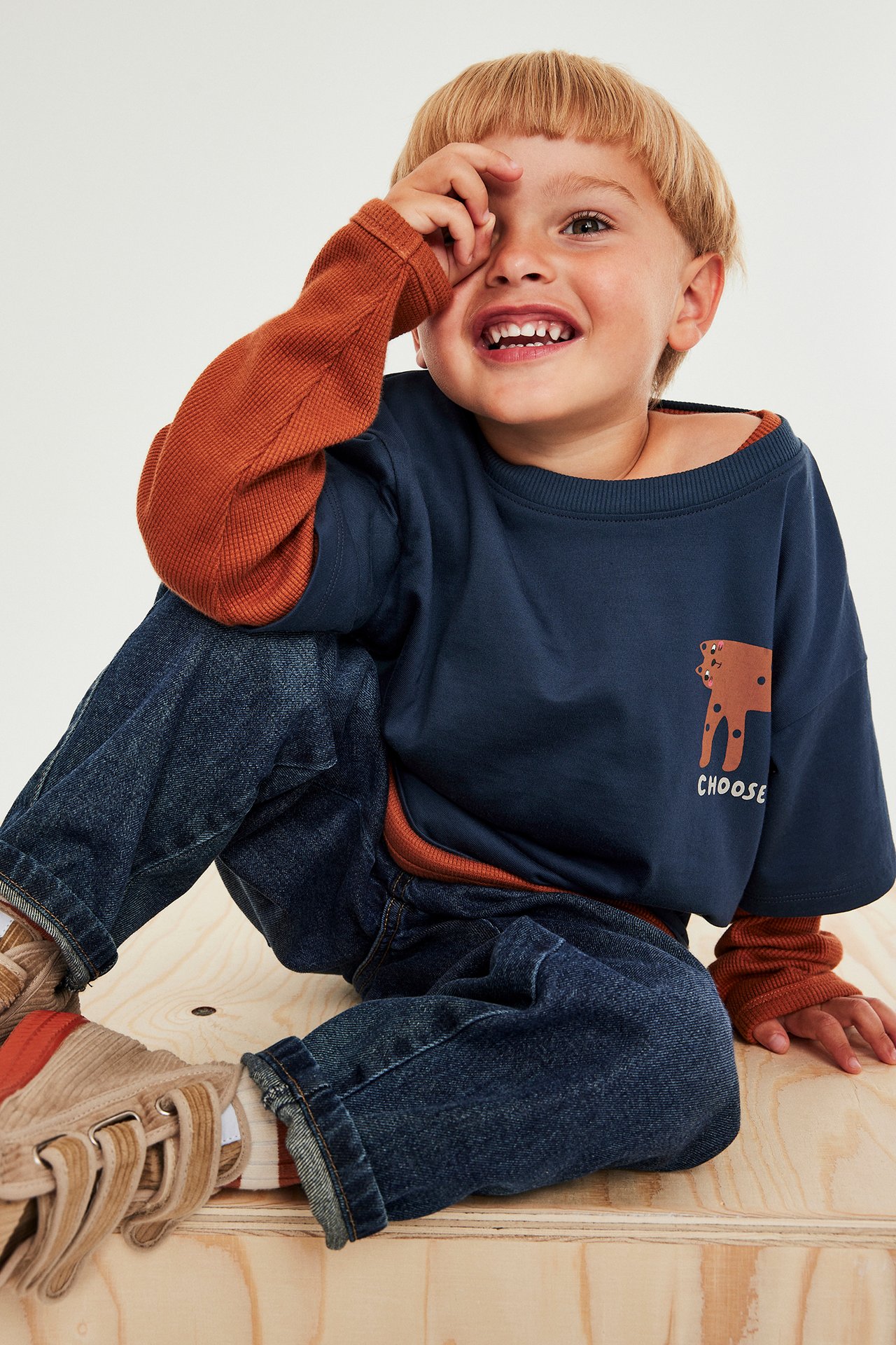 Barn i grå t-shirt med tryck, blå jeans och beige sneakers, sits på träbänk.