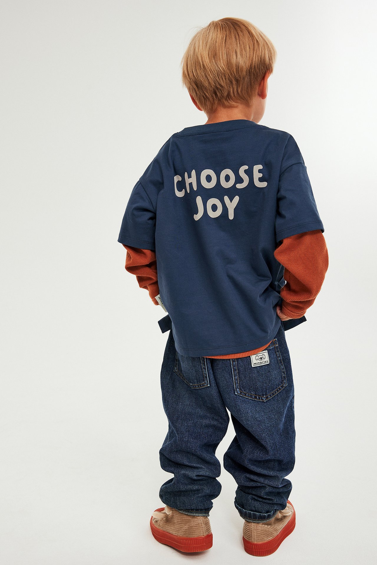 Barn med mörkblå T-shirt med tryck och modellering, bär jeans och fula skor, bakifrån.