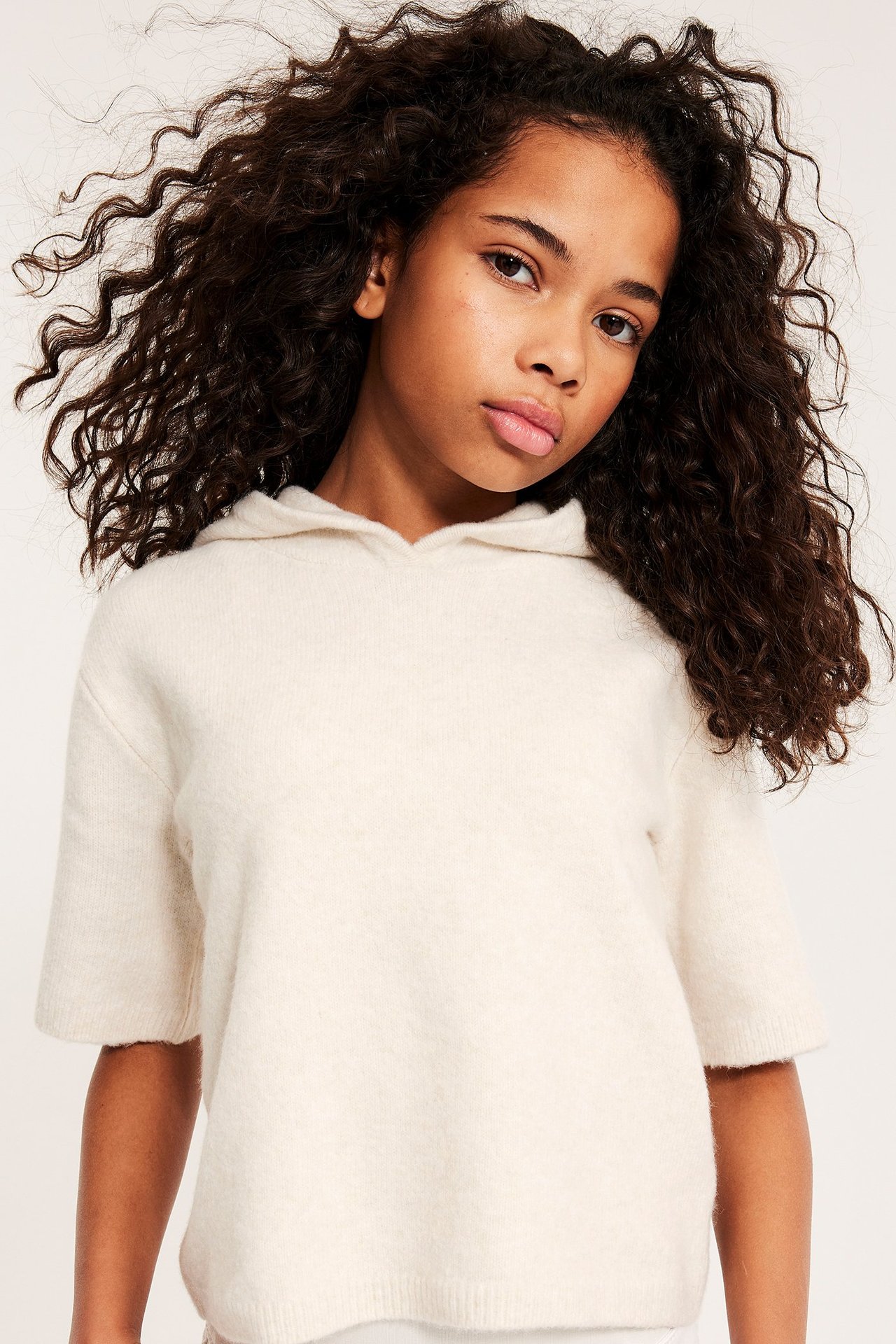 Framifrån, ett barn i en vit kortärmad stickad huvtröja över en vit topp.