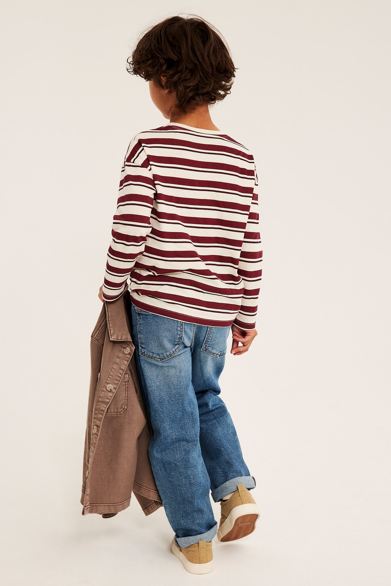 Bakifrån, barn i randig långärmad t-shirt, blå jeans, beige sneakers och håller en brun jacka.