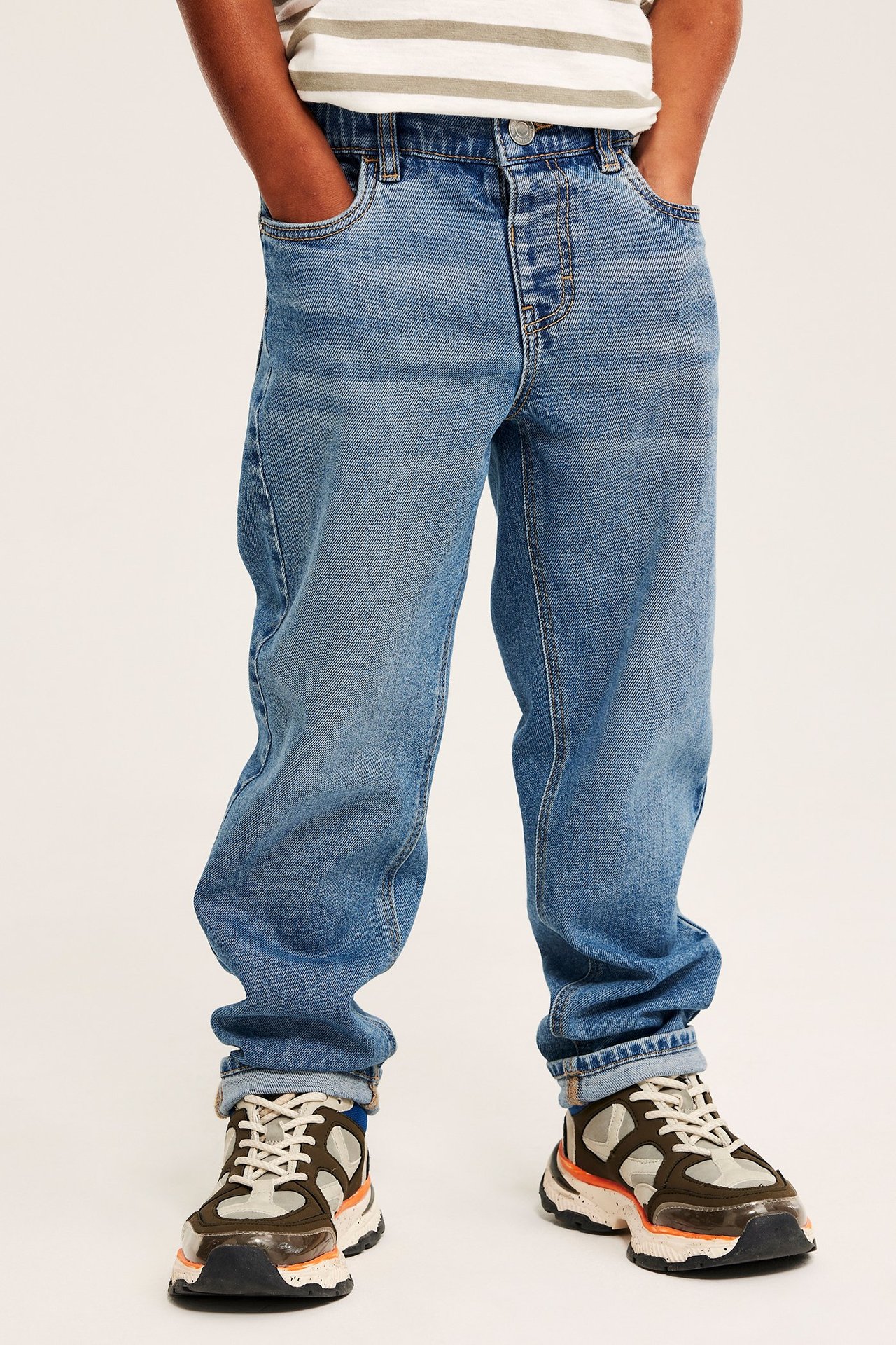 Frontvy av ett barn i blåa relaxed-fit denimjeans med uppvikta benslut, en randig t-shirt och grova sneakers.