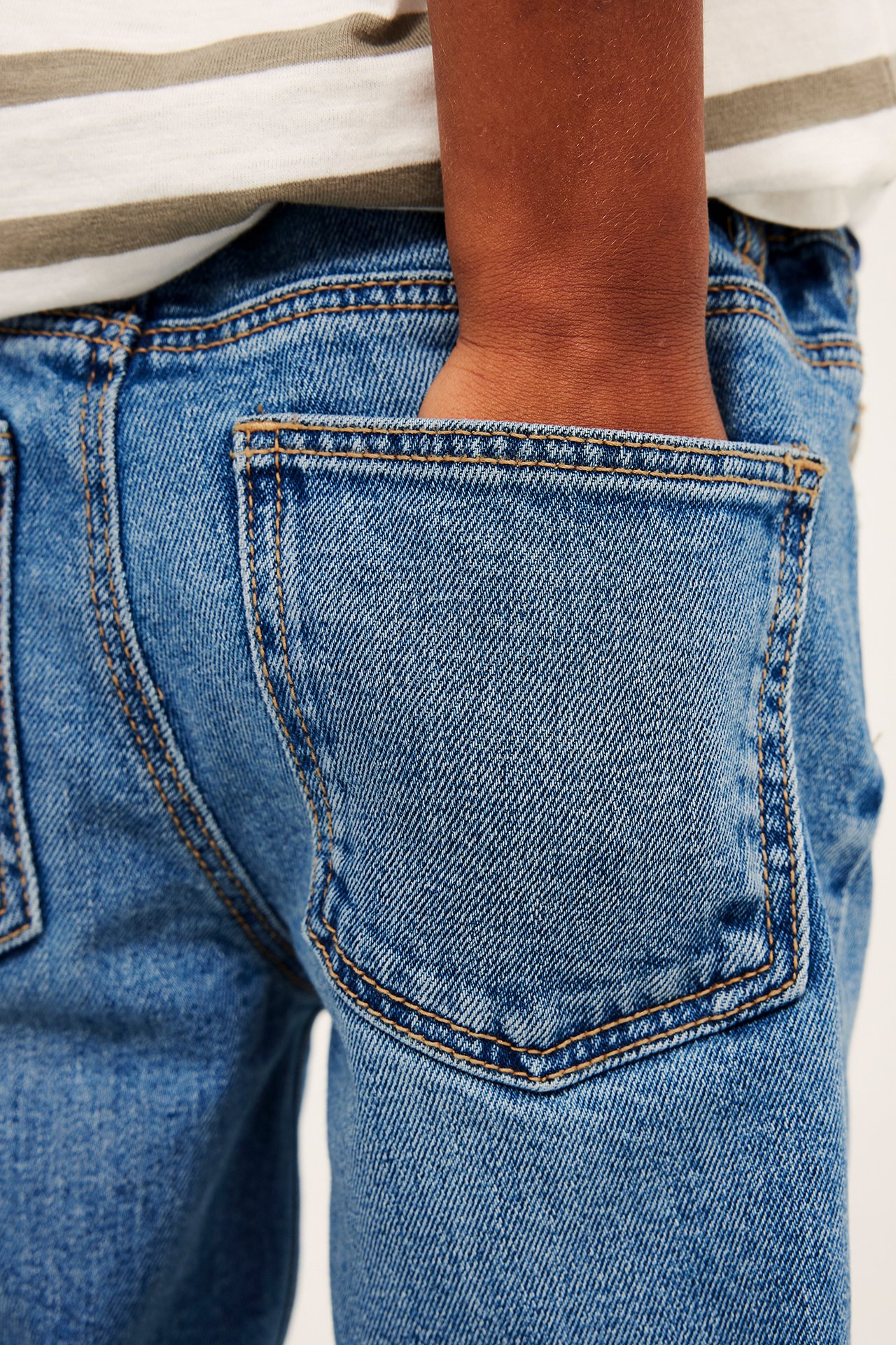 Närbild bakifrån: barn i lediga blå denimjeans, hand i bakfickan, över en randig t-shirt.