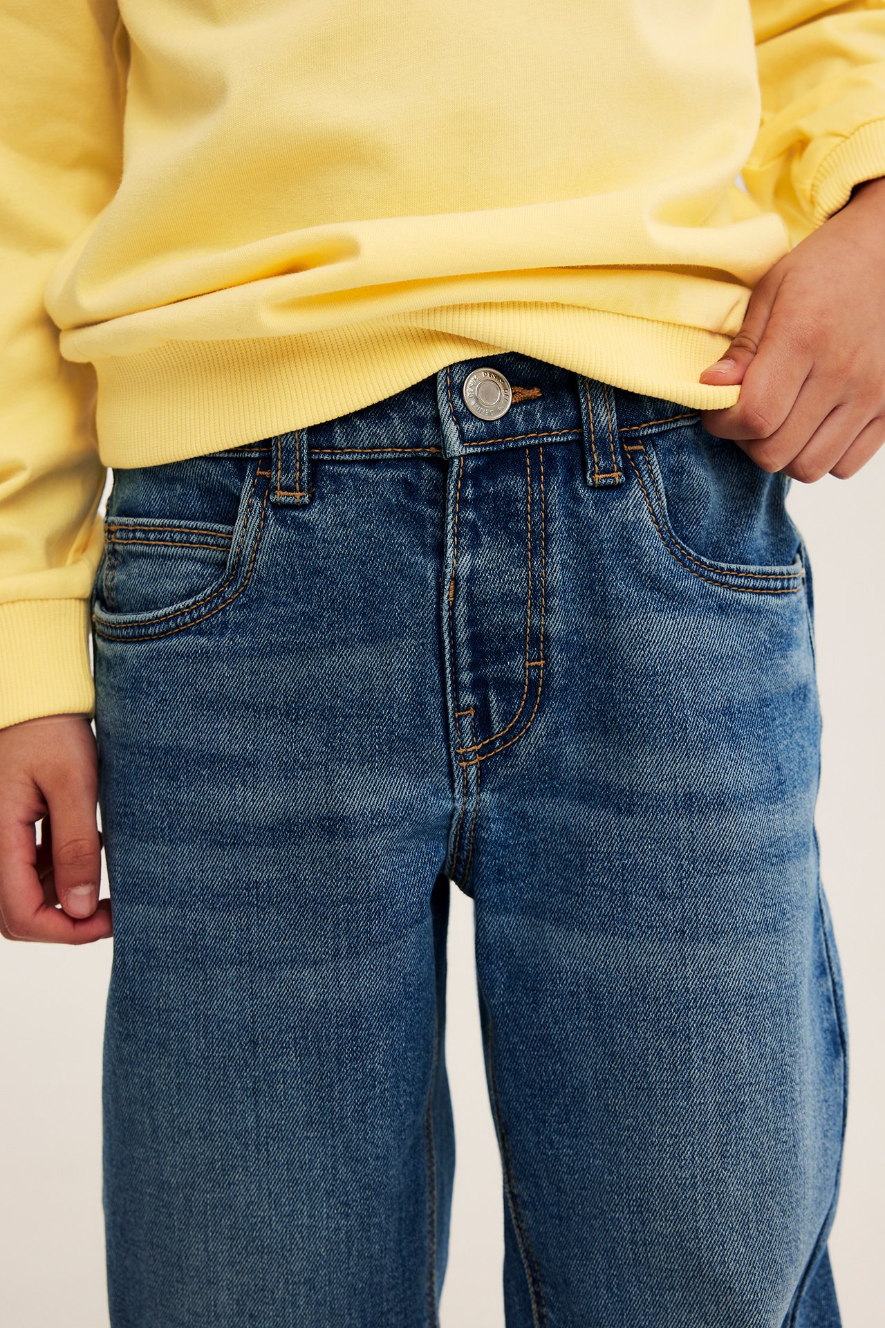 Frontvy av ett barn i avslappnade blå denimjeans med knapp och en gul sweatshirt.