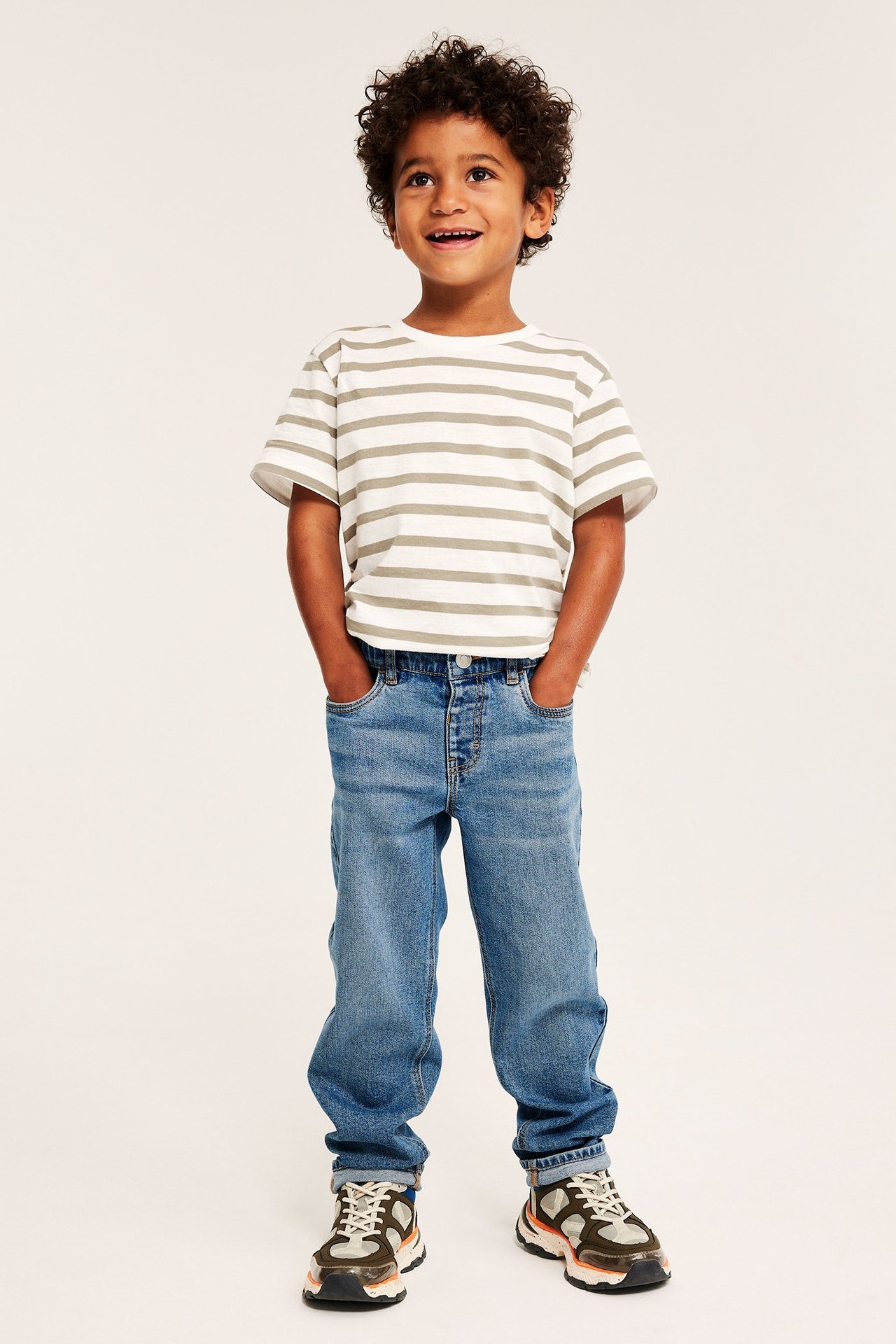 Widok z przodu uśmiechniętego dziecka w luźnych niebieskich dżinsach, pasiastym T-shircie i sneakersach.