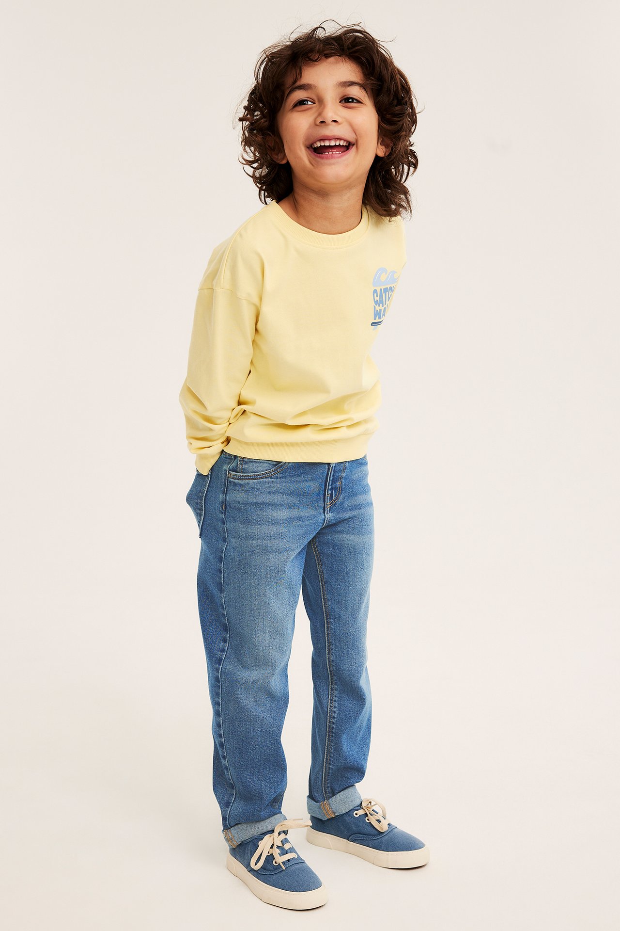 Forfra: et barn i blå relaxed denimjeans, en gul grafisk langermet genser og blå canvassko.