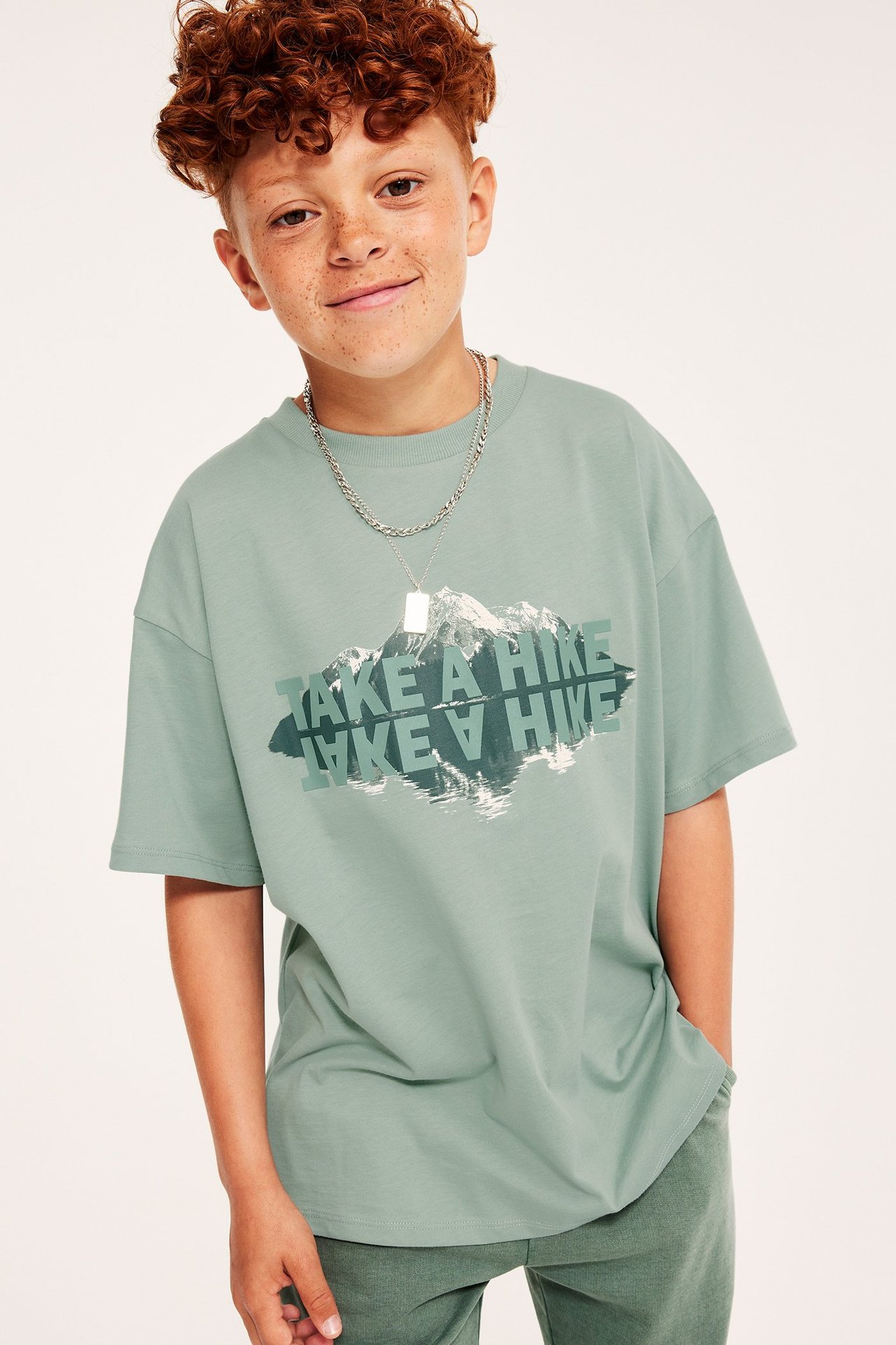 Frontvy: Barn bär ljusgrön oversized bomulls-T-shirt med bergstryck, gröna byxor och silverhalsband.