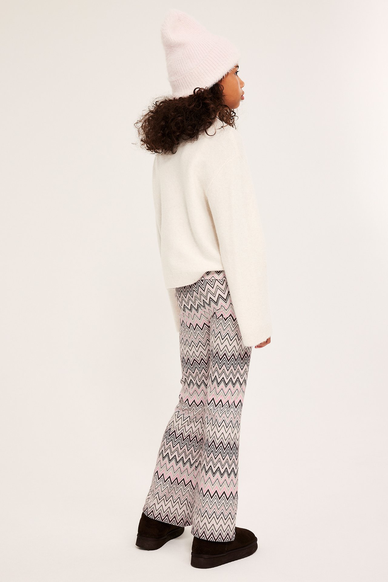 Sideprofil av et barn i mønstrede beige slengleggings, kremfarget genser, lys rosa lue og svarte støvler.