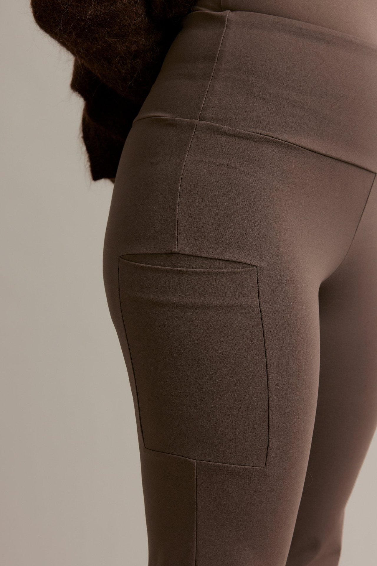 Lähikuva naisesta tummanruskeissa korkeavyötäröisissä bootcut treenileggingseissä, joissa sivutasku lahkeessa.