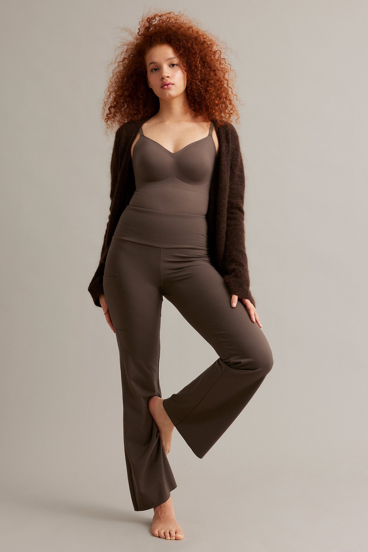 Edestäpäin: Nainen tummanruskeissa korkeavyötäröisissä bootcut-treenitrikoissa, joissa tasku, yhteensopiva toppi, pörröinen neuletakki.