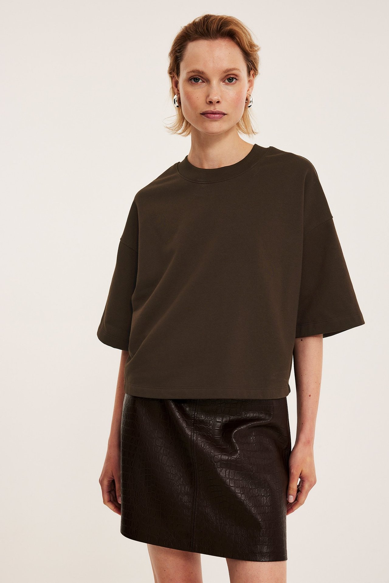 Widok z przodu: Kobieta w ciemnobrązowej bluzie z krótkim rękawem, ciemnobrązowej spódniczce mini z krokodylim wzorem, srebrnych kolczykach.