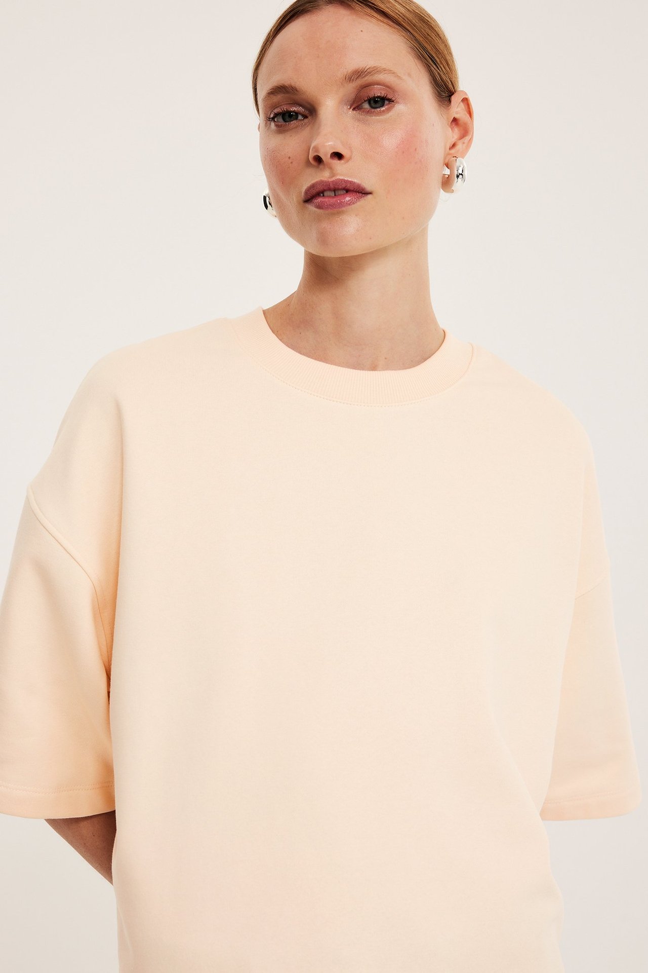 Lähikuva edestä naisesta, jolla on väljä vaaleanbeige lyhythihainen collegepaita ja hopeiset rengaskorvakorut.