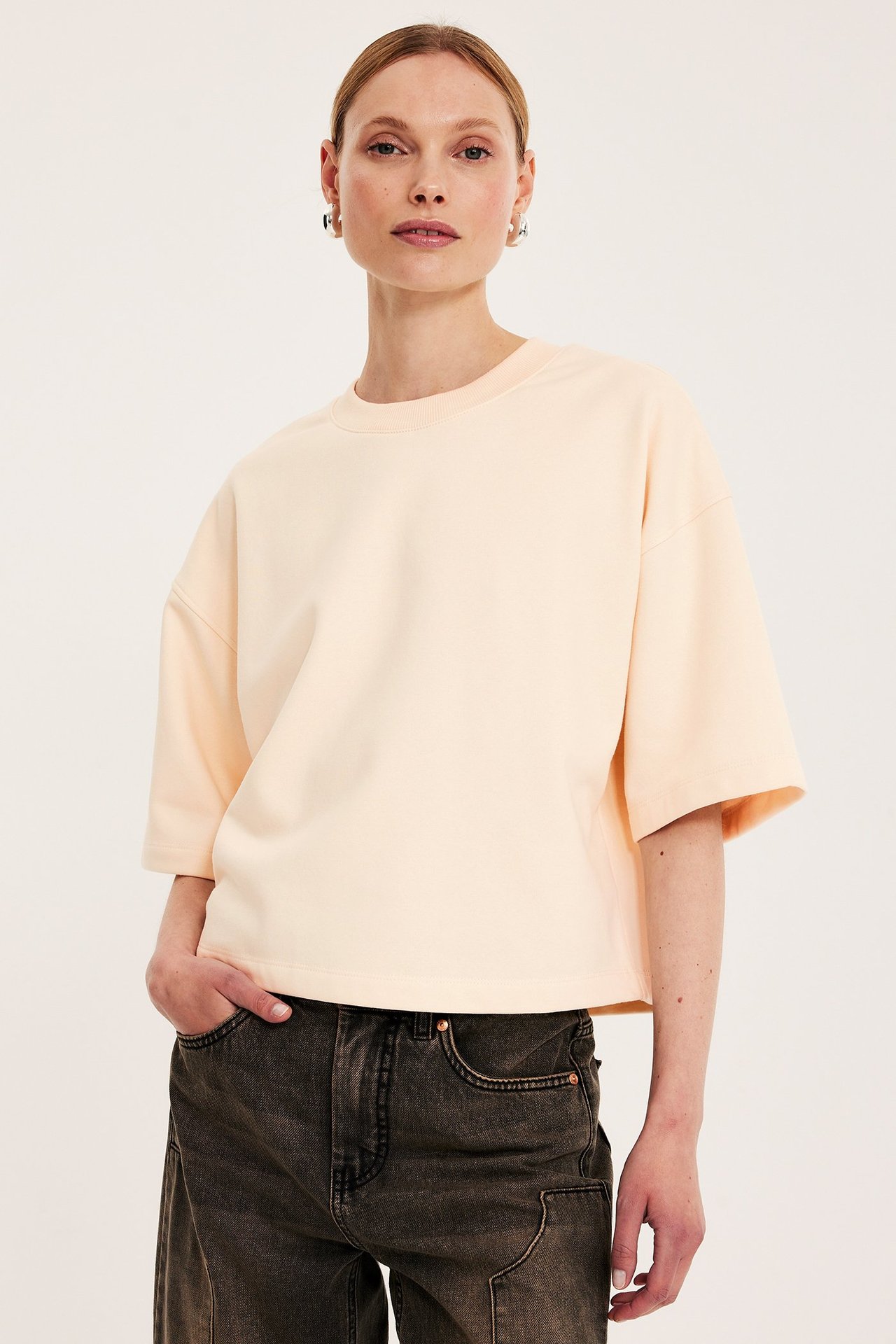 Widok z przodu: Kobieta w jasnobeżowej bluzie z krótkim rękawem, ciemnoszarych dżinsach i srebrnych kolczykach kołach.