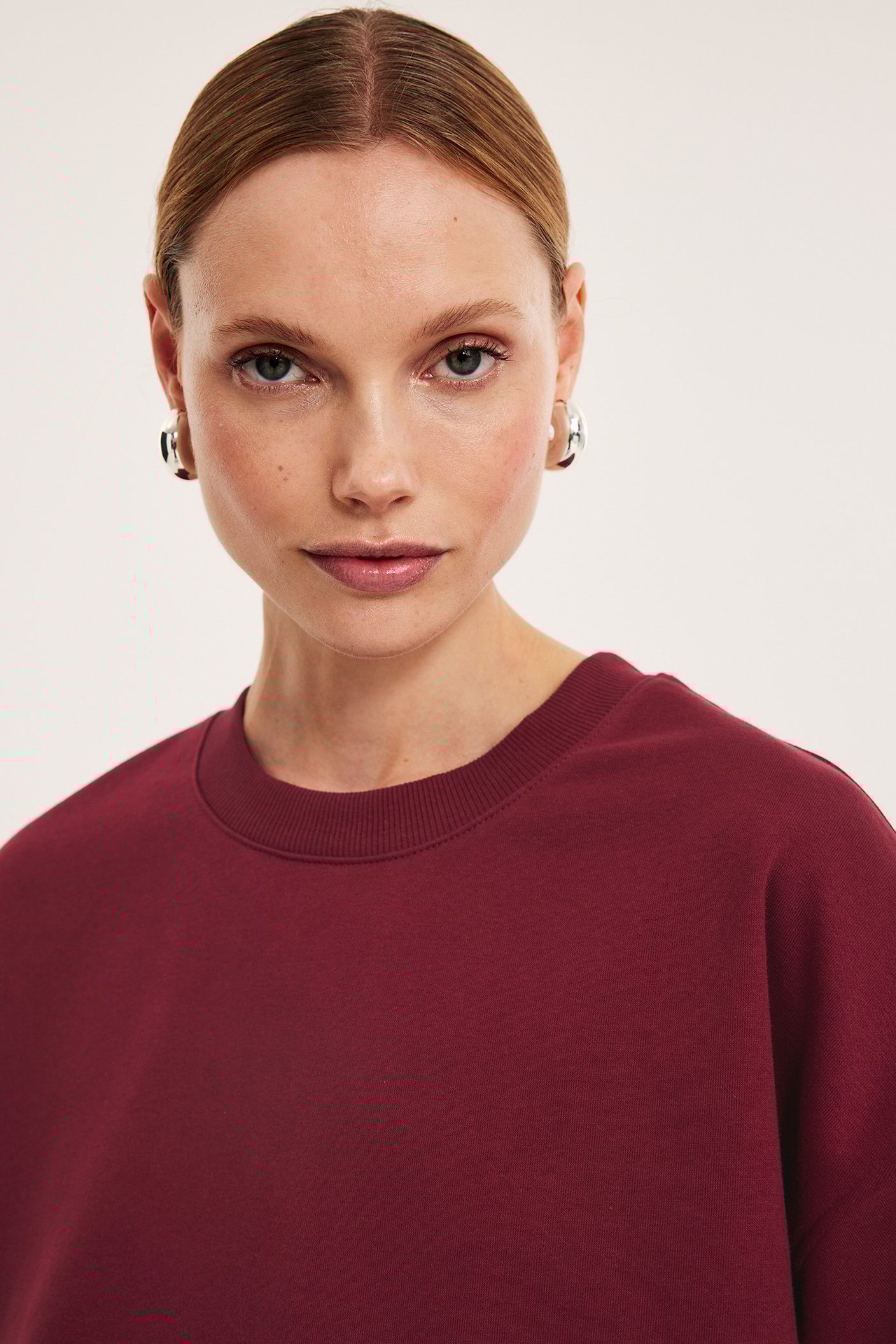 Lähikuva naisesta yllään tummanpunainen lyhythihainen collegepaita ja hopeiset rengaskorvakorut.