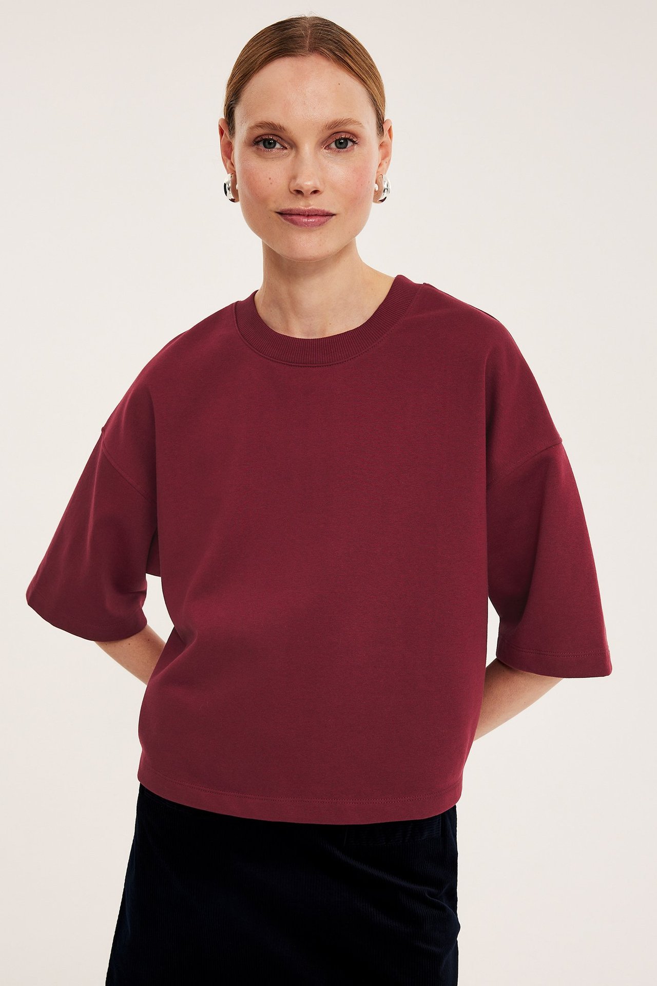 Widok z przodu: kobieta w ciemnoczerwonej bluzie z krótkim rękawem, granatowej spódnicy i srebrnych kolczykach.