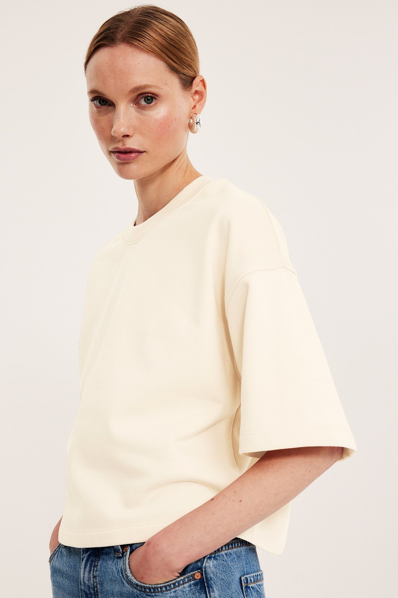 Widok z boku kobiety w kremowej, luźnej bluzie z krótkim rękawem, niebieskich dżinsach i srebrnych kolczykach.