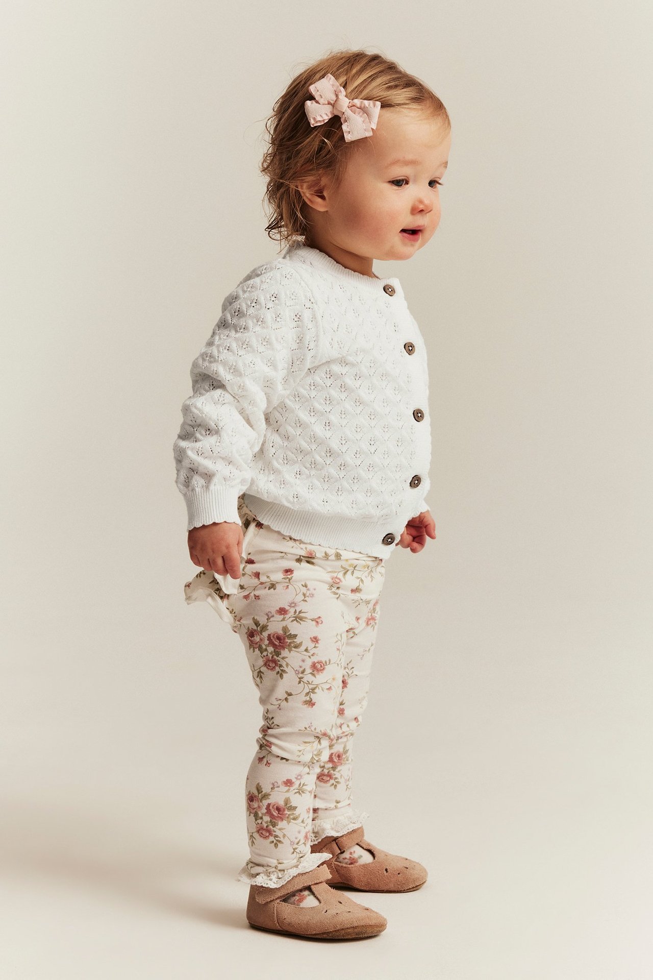 Sidebilde av en baby i en mønstret off-white bomullskofte med knapper, blomstrete leggings og rosa sløyfe.