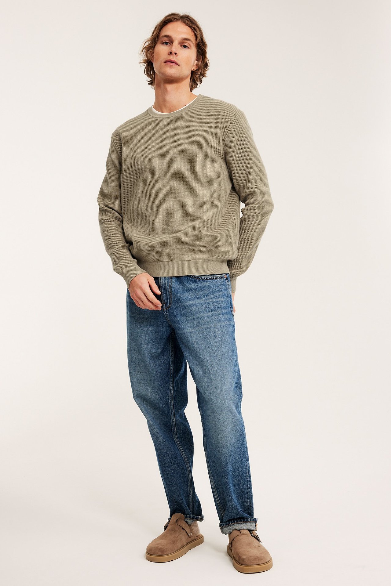 Edestäpäin kuvattu mies beigessä neuleessa, sinisissä farkuissa ja beigenvärisissä slip-on-kengissä.