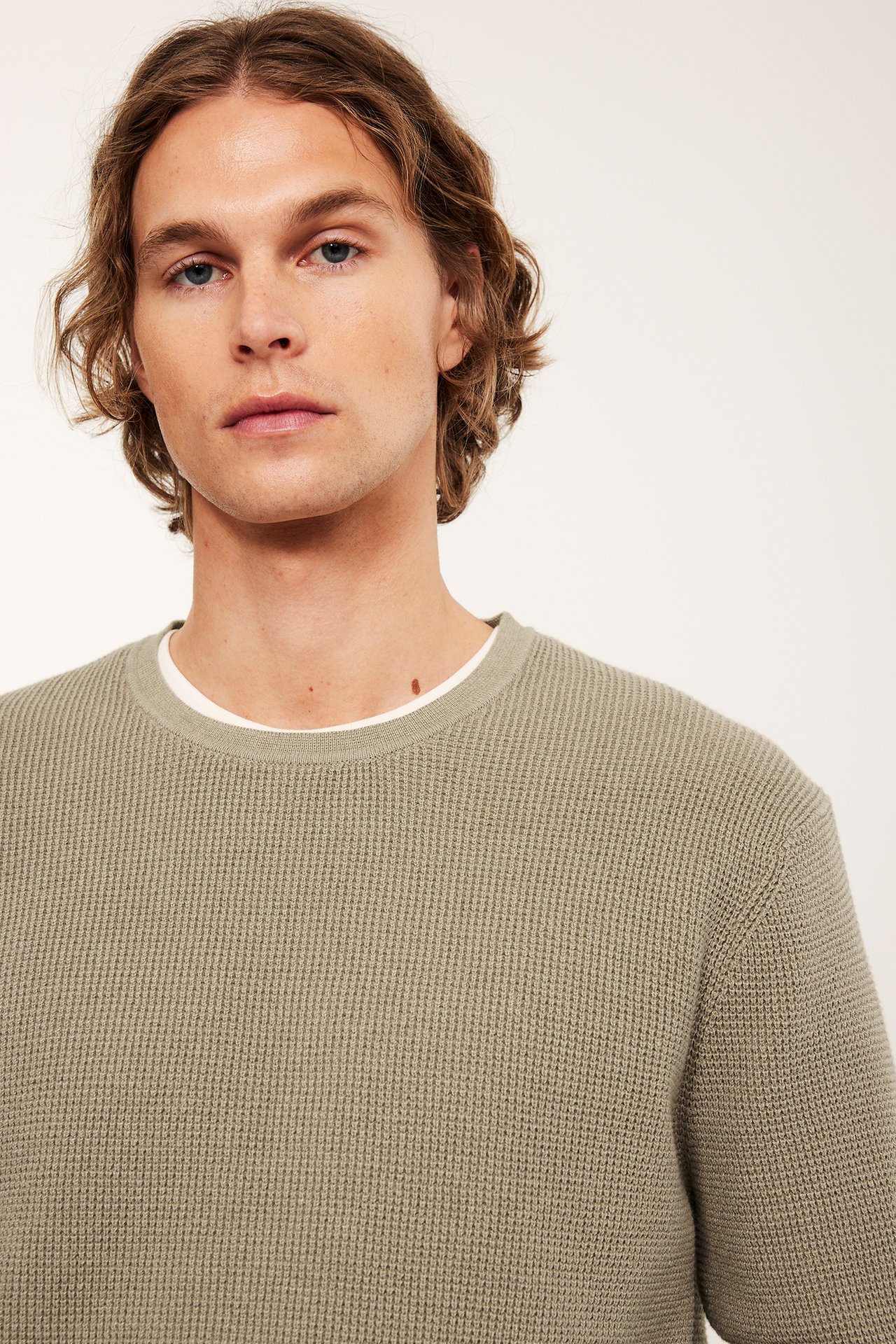 Lähikuva edestä miehestä, yllään beigemeleerattu neulepusero valkoisen t-paidan päällä.
