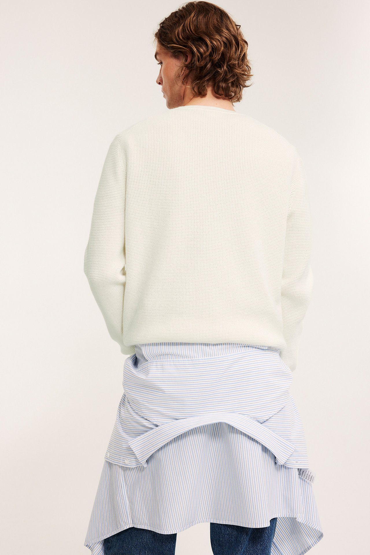 Miehen takaapäin: offwhite melange-neulepusero, sinivalkoraidallinen paita sidottuna vyötäröllä.