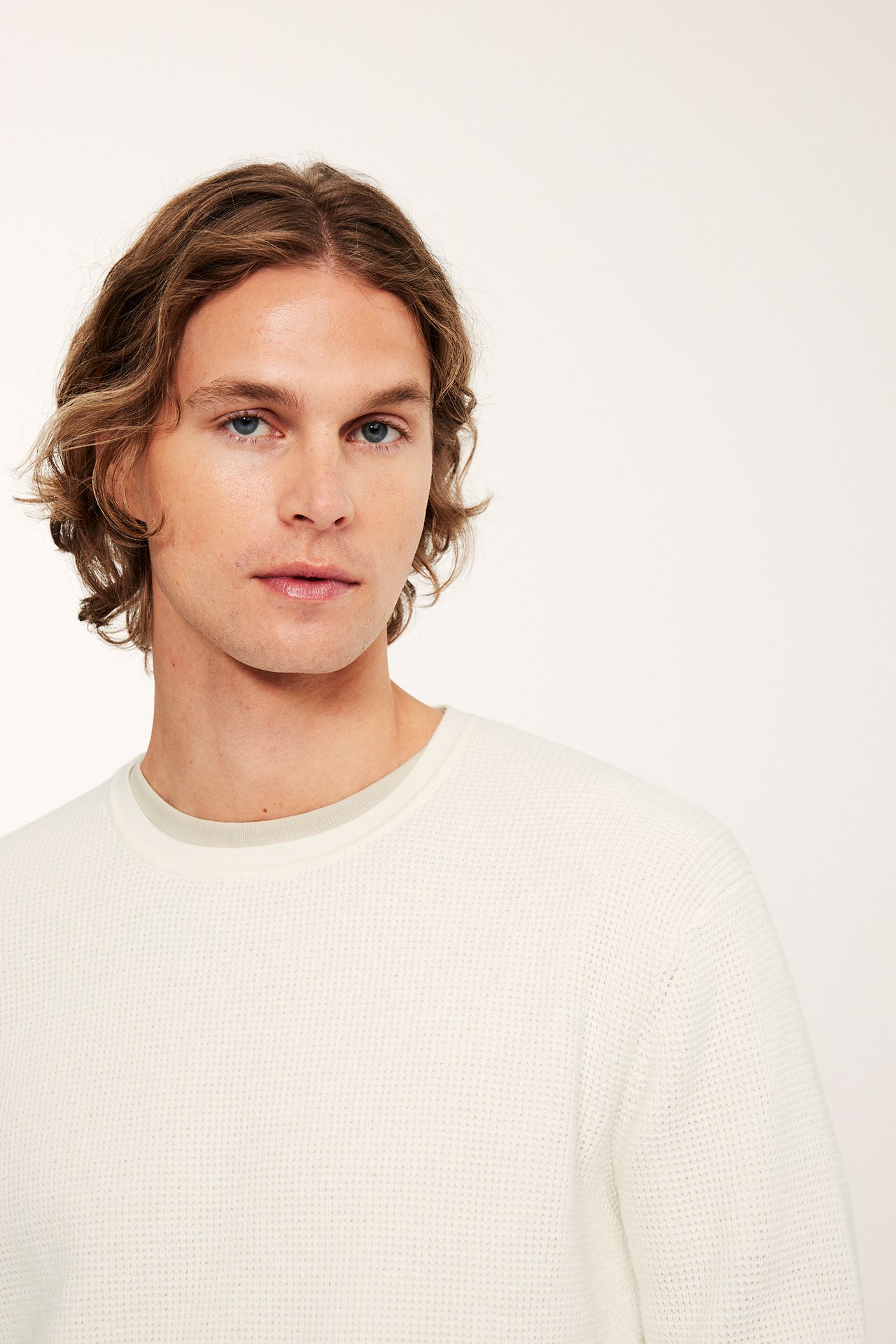 Lähikuva miehestä, yllään luonnonvalkoinen meleerattu neulepusero vaaleanharmaana t-paidan päällä.