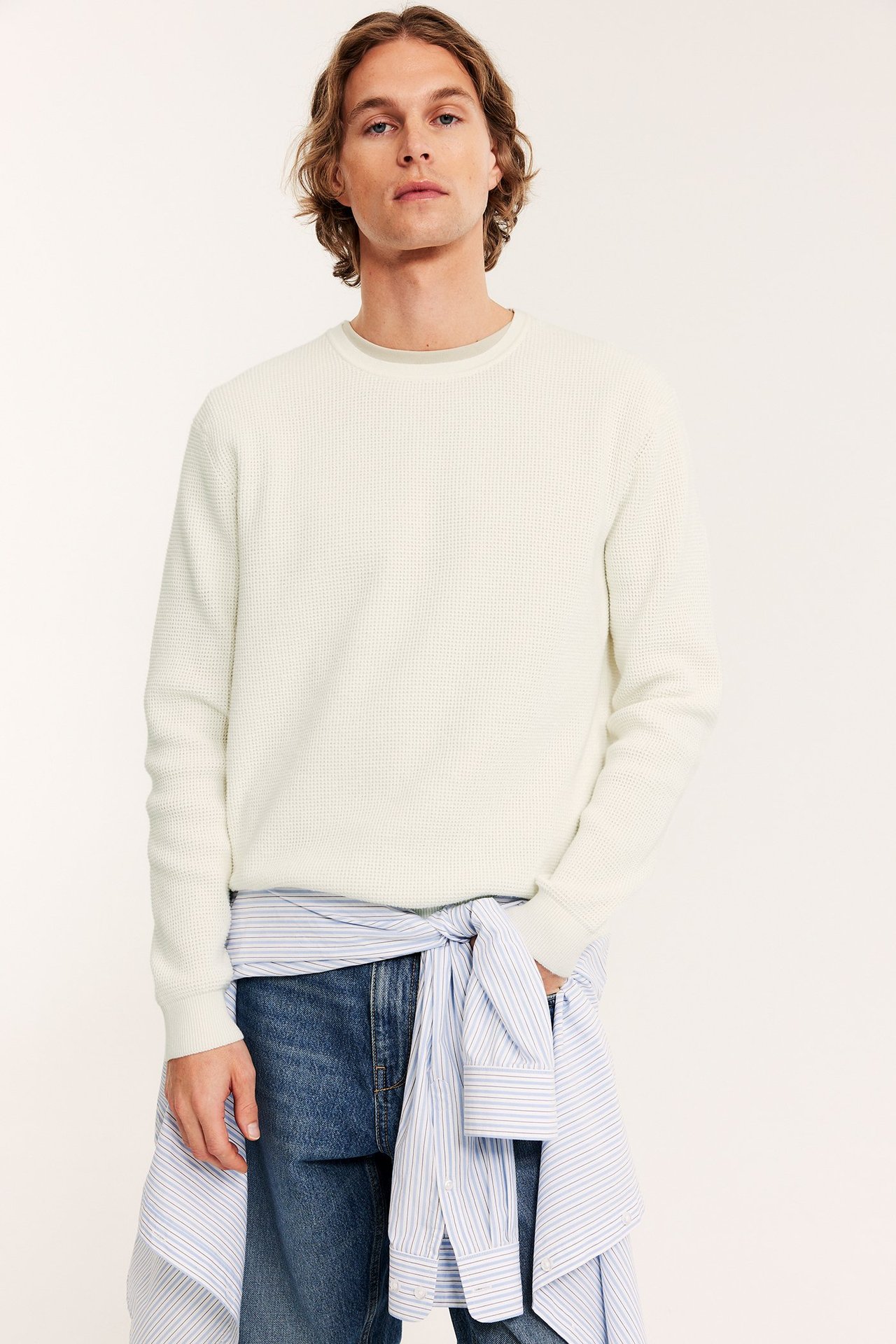 Front: Mann i offwhite melert strikket genser, blå jeans, med stripet skjorte knytt rundt midjen.