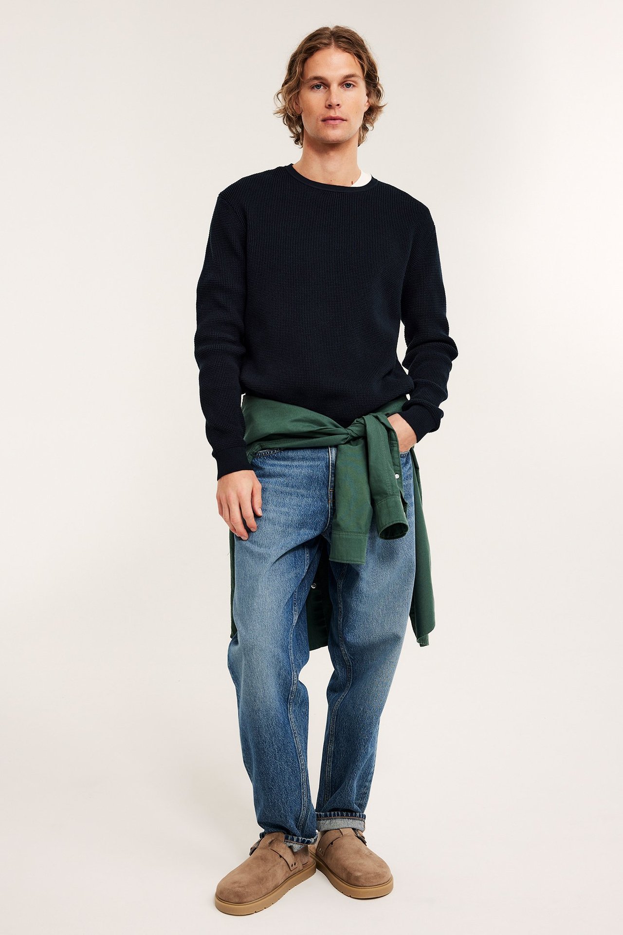 Mann i mørkeblå strikkegenser, lyseblå jeans og beige tøfler. Grønn skjorte knytt rundt midjen, forfra.