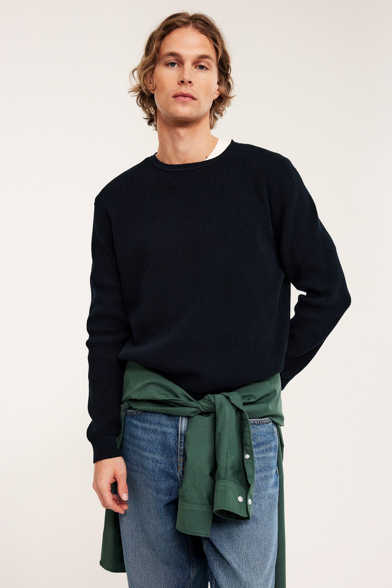 Frontutsikt: Mann i mørkeblå vaffelstrikket genser, lyseblå jeans, grønn skjorte rundt midjen.