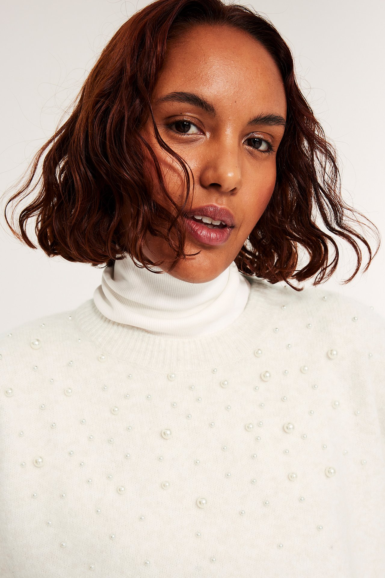 Kvinna i vit stickad tröja med pärlor, tavelvinkel. Fjädrande hår, turtleneck.