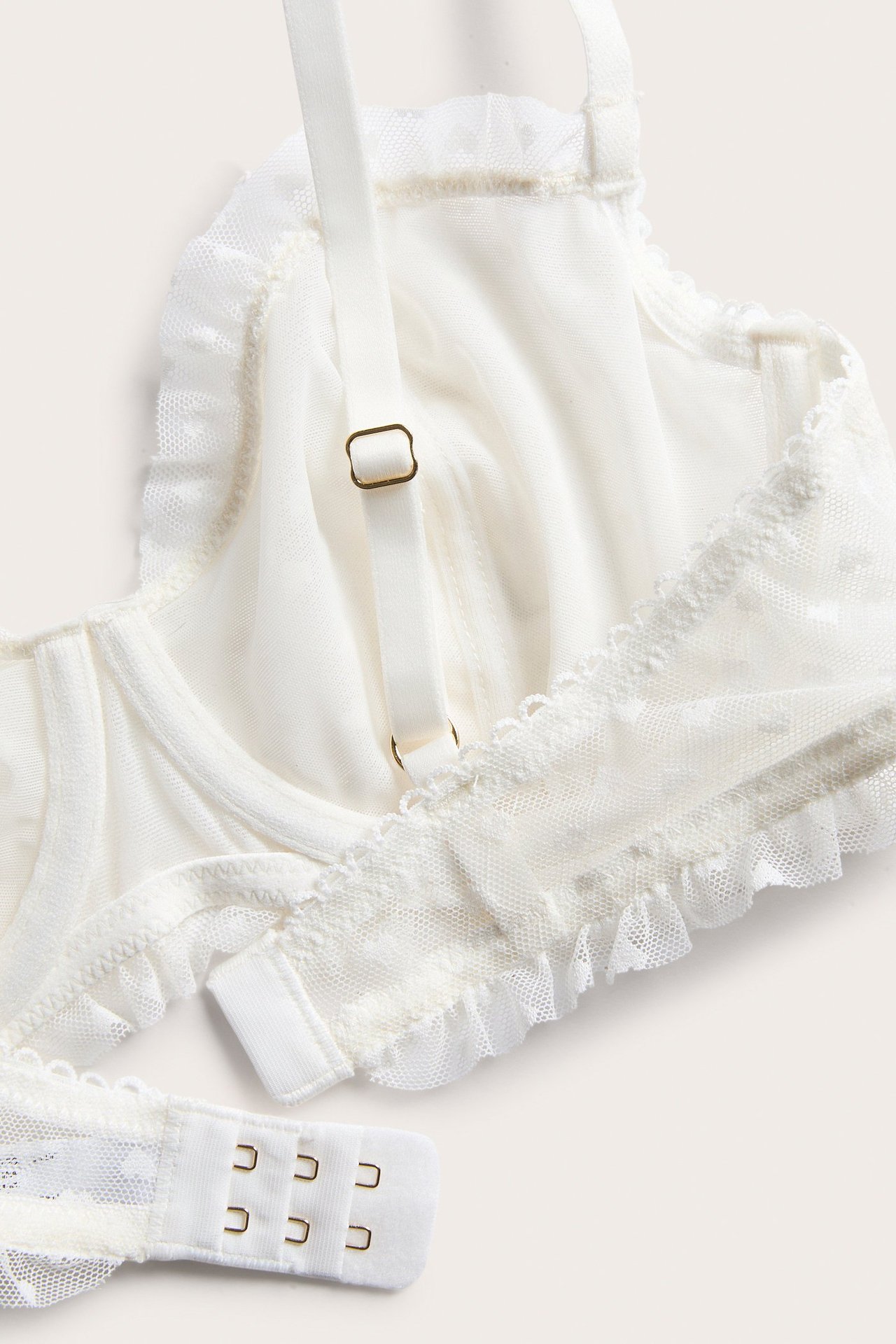 Zbliżenie biustonosza z koronki off-white w serca, falbany, regulowane ramiączka i zapięcie na haftki.