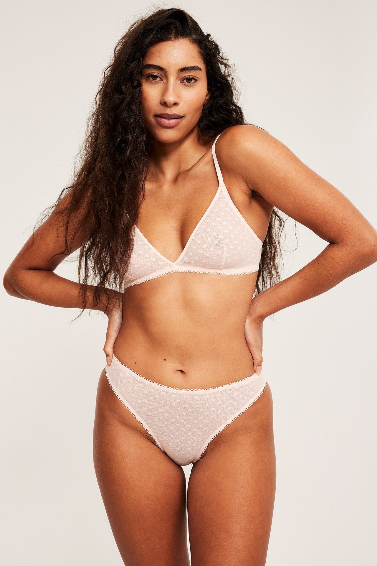 Frontbilde av kvinne i lyserosa, ufôret mesh-bralette med hjertemønster og matchende truser.
