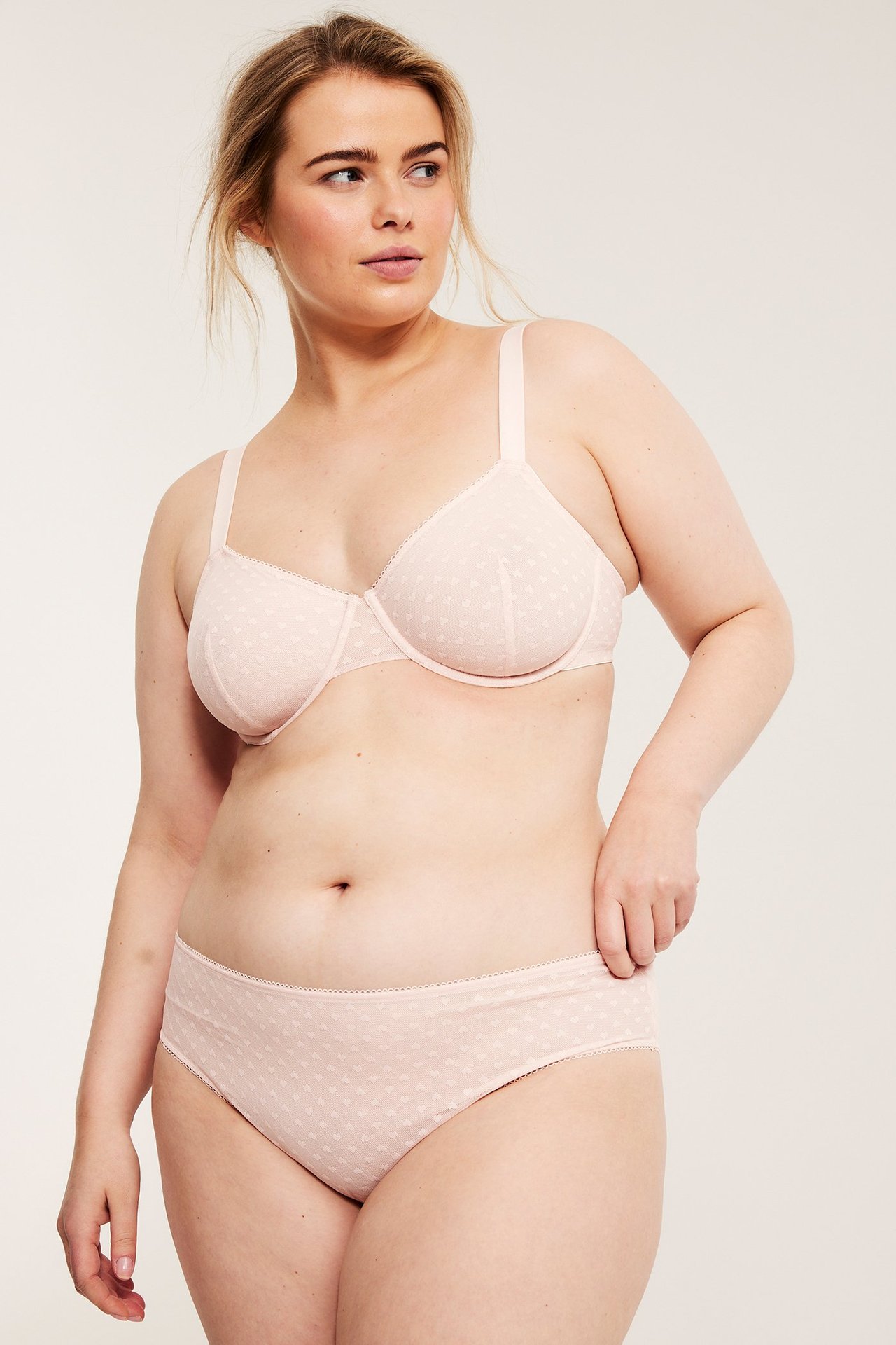 Frontbilde av en kvinne i en lys rosa, uvattert spile-BH og matchende truse med hjertemønster.