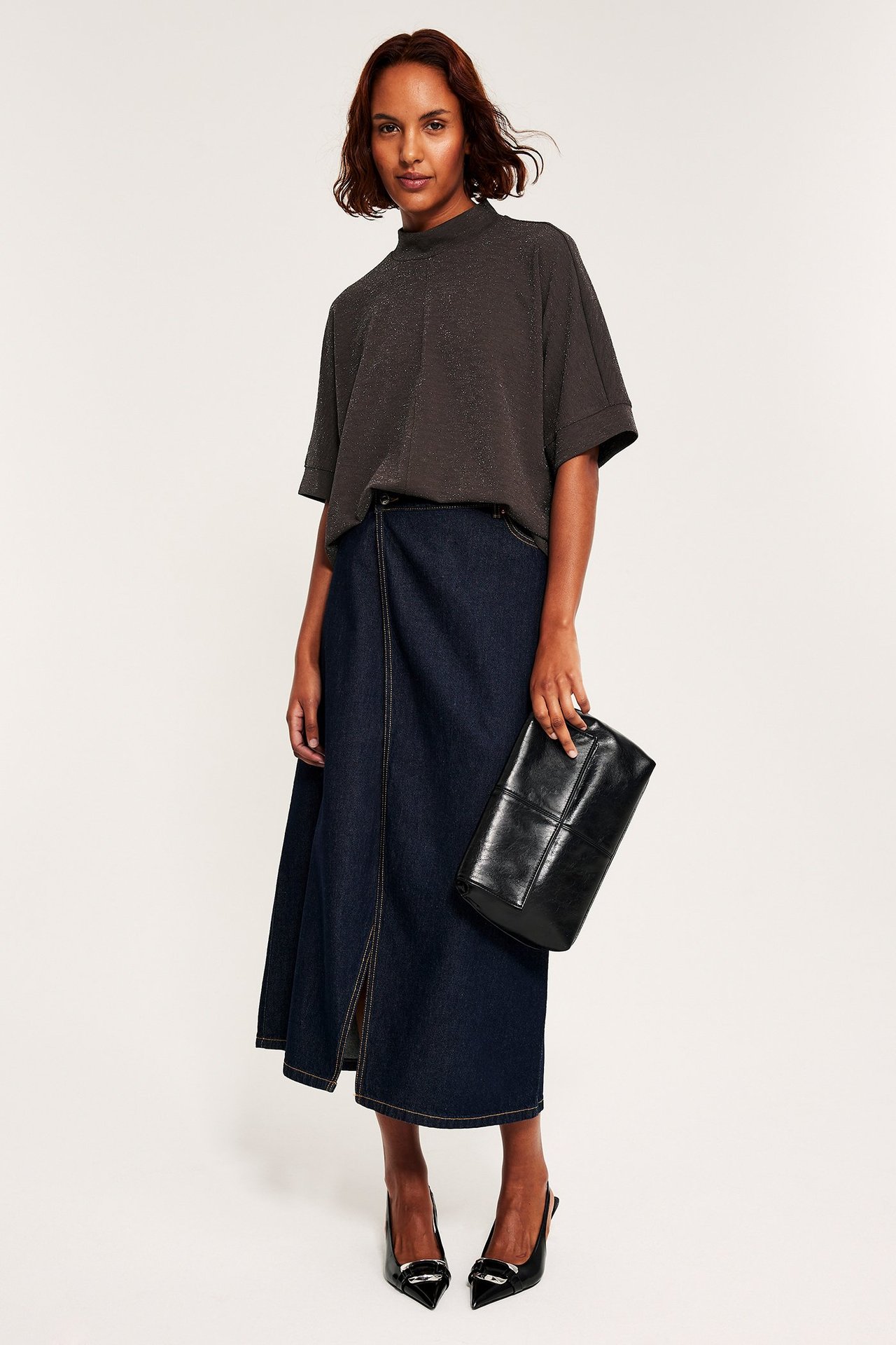 Kvinna i mörkbrun glittrig löst sittande topp, blå jeansomlottkjol, svarta klackar, håller svart clutch.