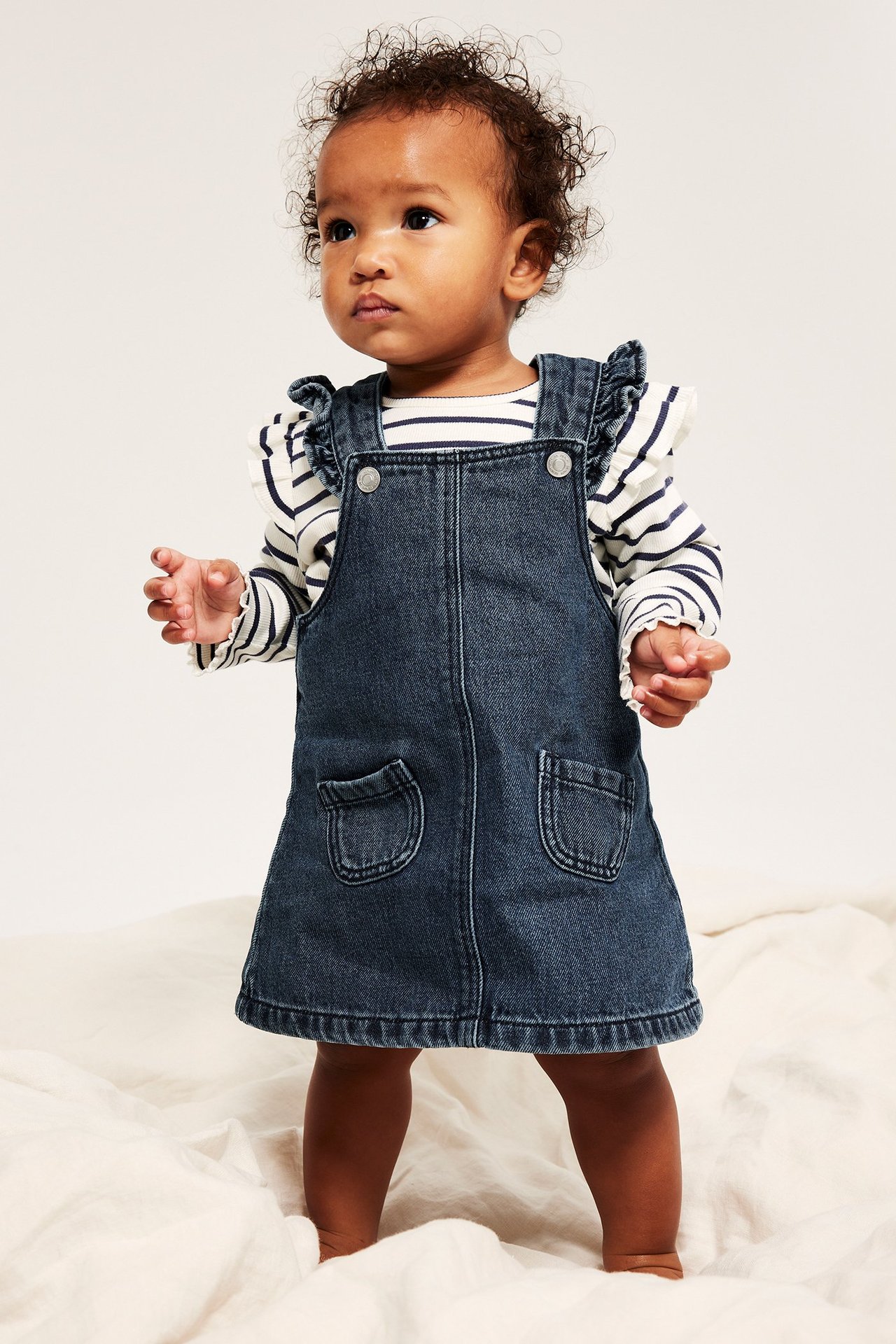 Edestäpäin vauva tummansinisessä farkkuolkahameessa, jossa röyhelöt ja taskut, raidallisen pitkähihaisen paidan päällä.