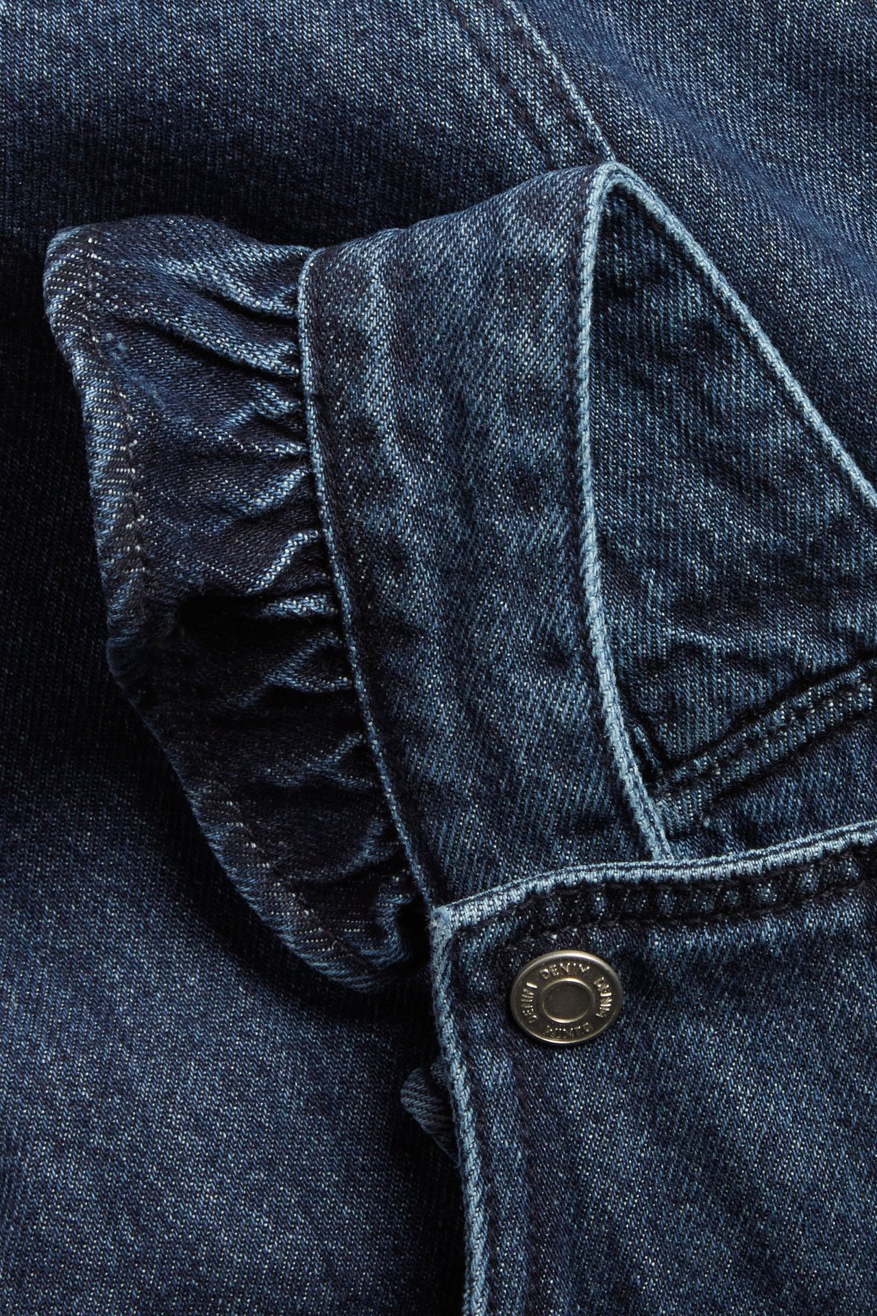 Lähikuva vauvan denim-mekosta, jossa on röyhelö olkapäällä ja hopeinen neppari.