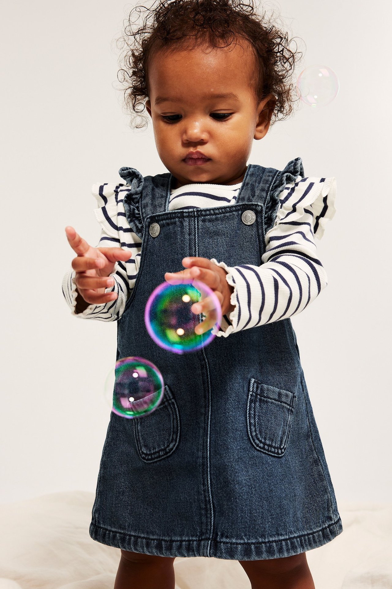 Edestä: Vauva deniminsinisessä röyhelömekossa taskuilla valkoisen raidallisen paidan päällä.