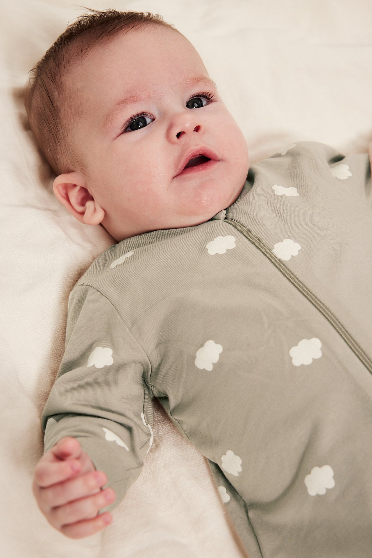 Baby i offwhite romper med molnprint och dragkedja, liggande på kudde, frontvy.