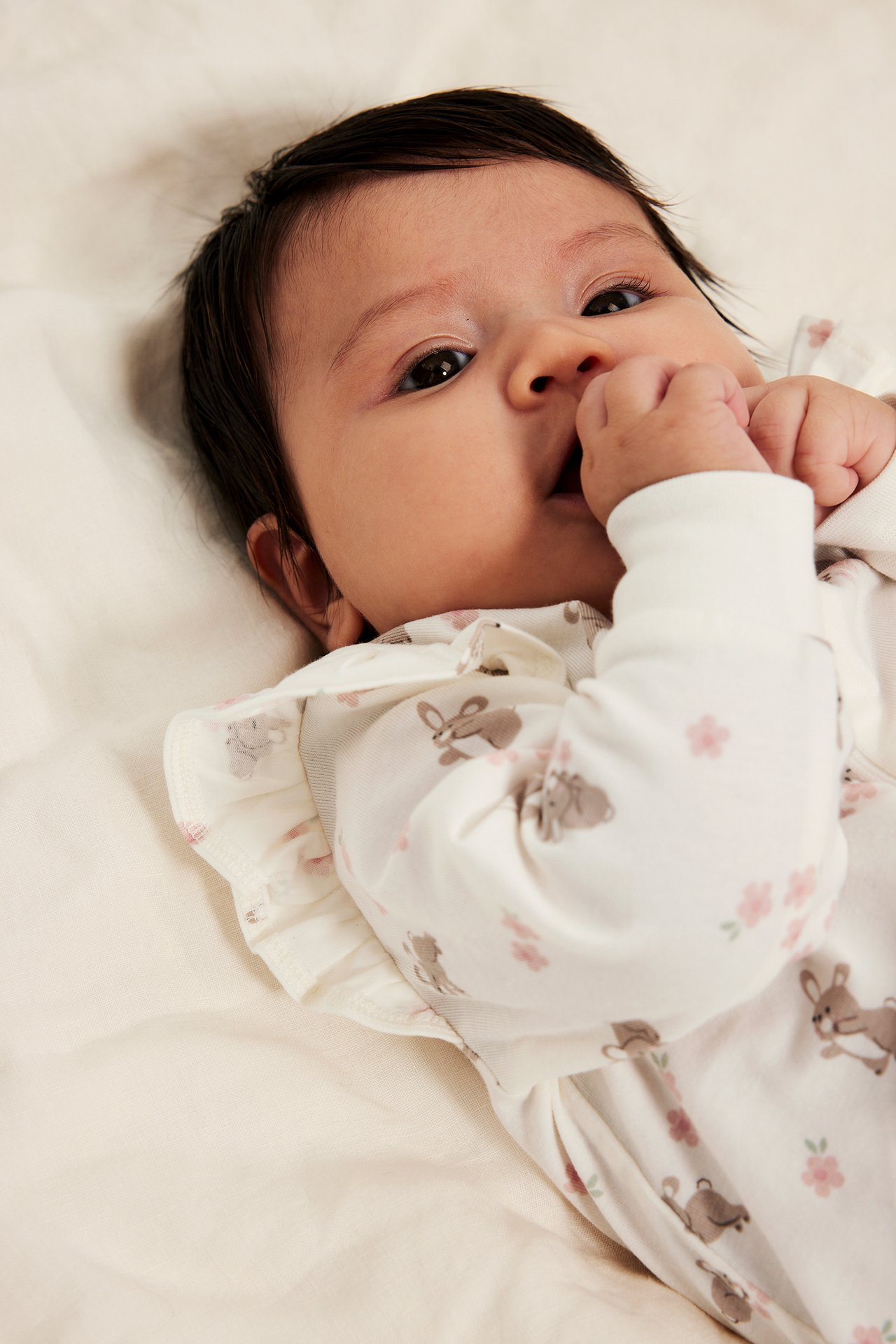 Nærbilde forfra av baby i off-white mønstret bomullspyjamas med volanger, kaniner og rosa blomster.