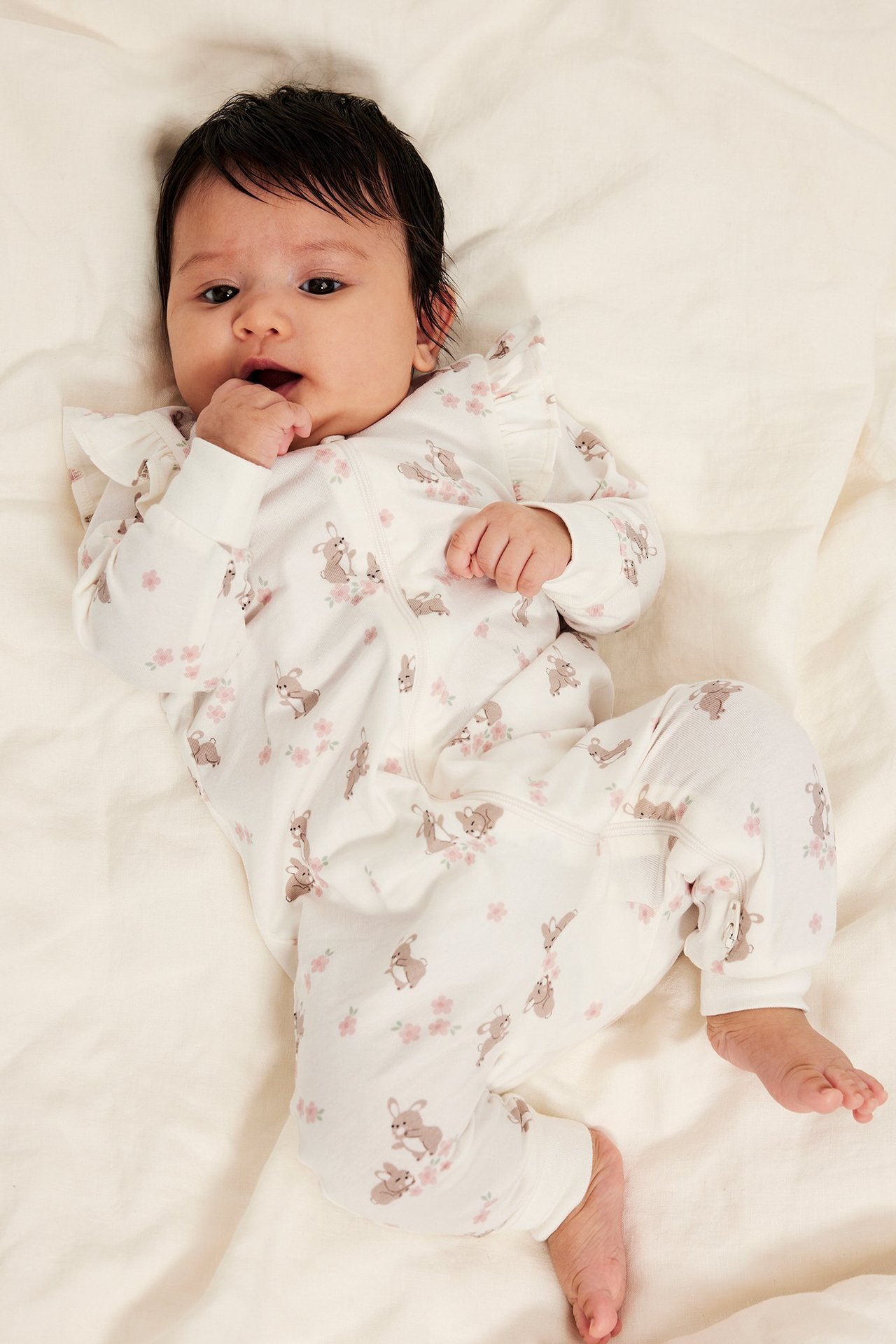 Widok z góry: Niemowlę w kremowej piżamie z falbanami, wzorem królików i różowych kwiatów, z zamkiem z przodu.