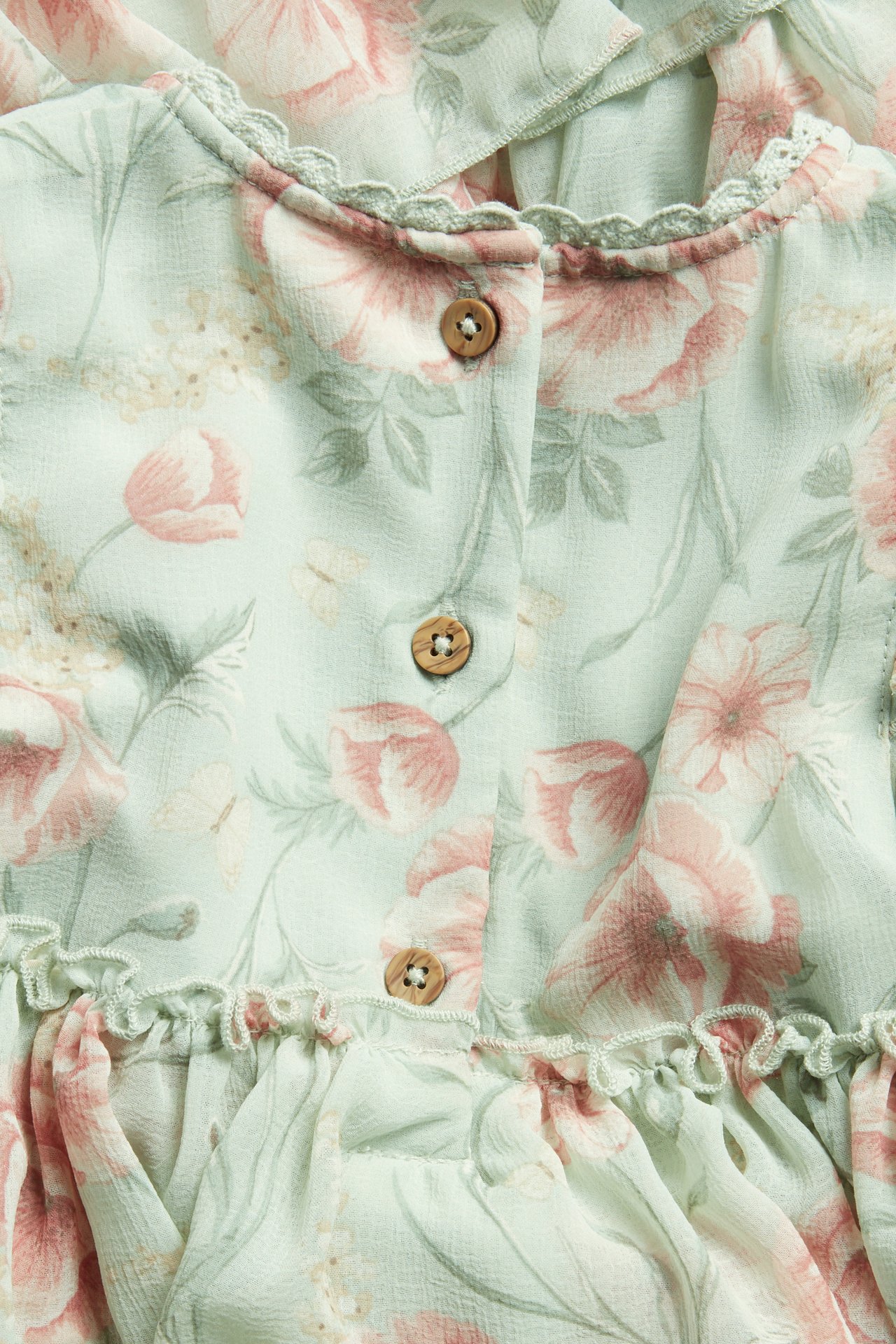Närbild bakifrån på en ljusgrön blommig chiffongklänning med volanger, spetskant och träknappar.