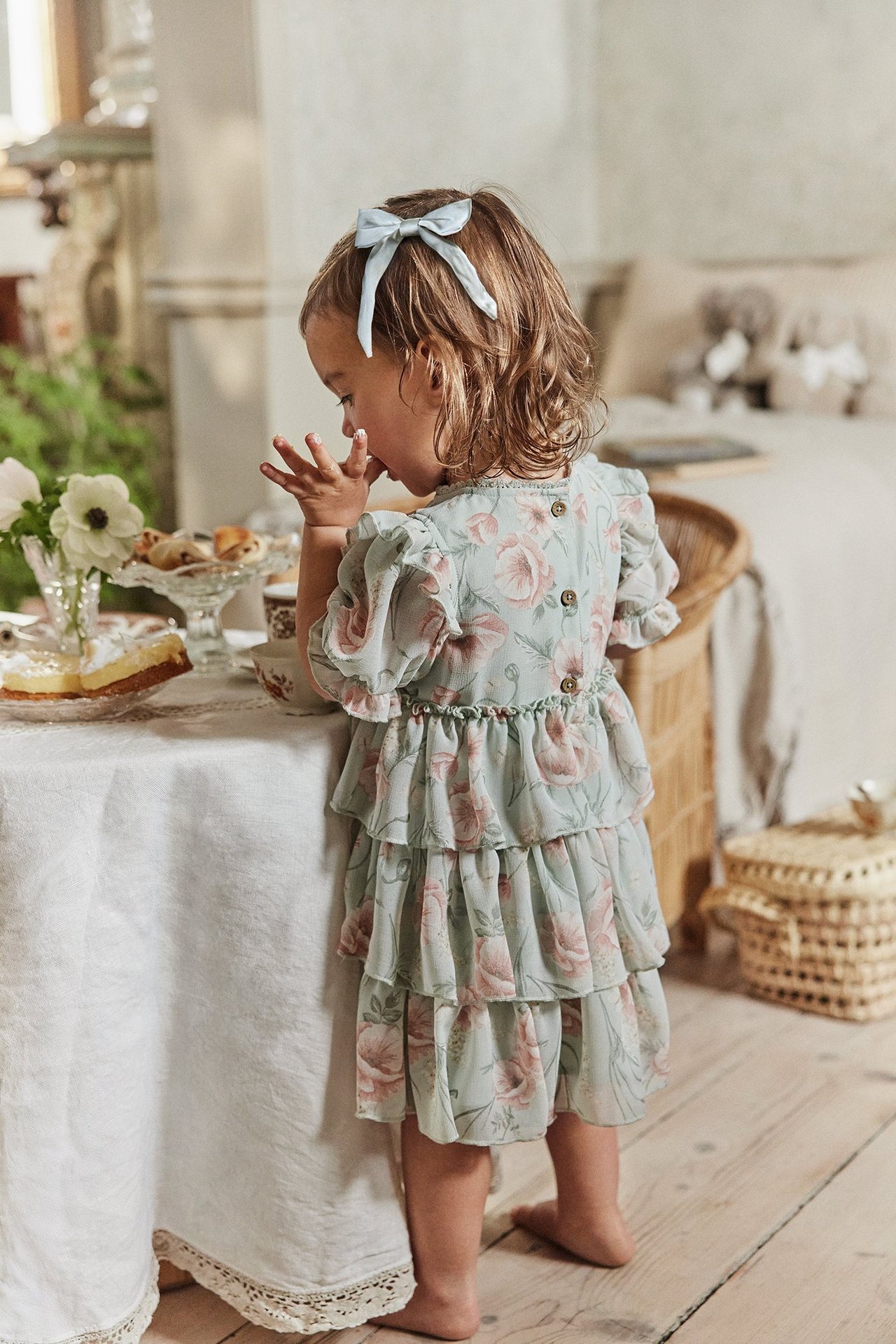 Widok od tyłu dziecka w jasnozielonej kwiecistej szyfonowej sukience z falbanami i niebieską kokardką.