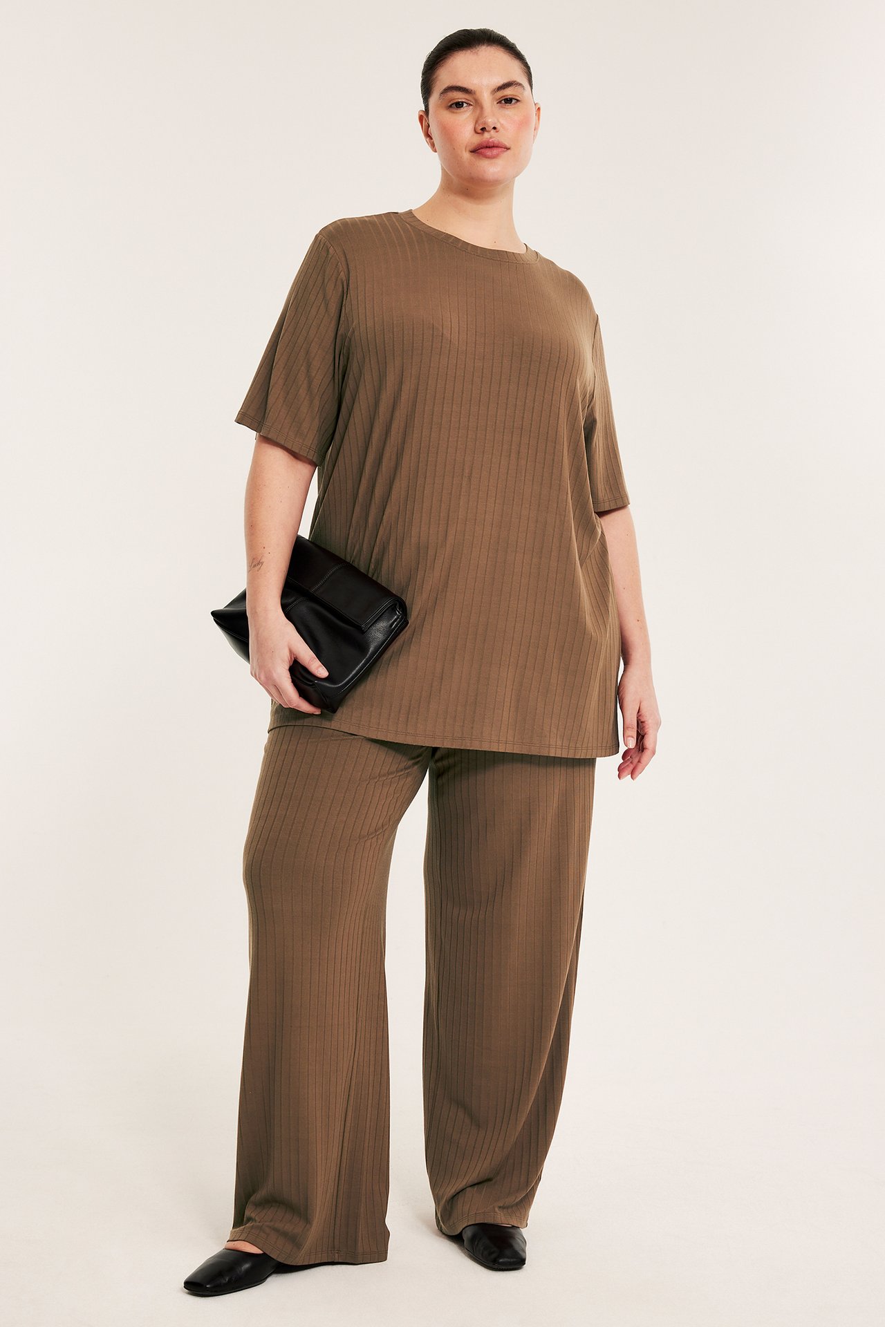 Frontvy av en kvinna i lång, brun, ribbad oversized t-shirt, matchande byxor, håller svart clutchväska.