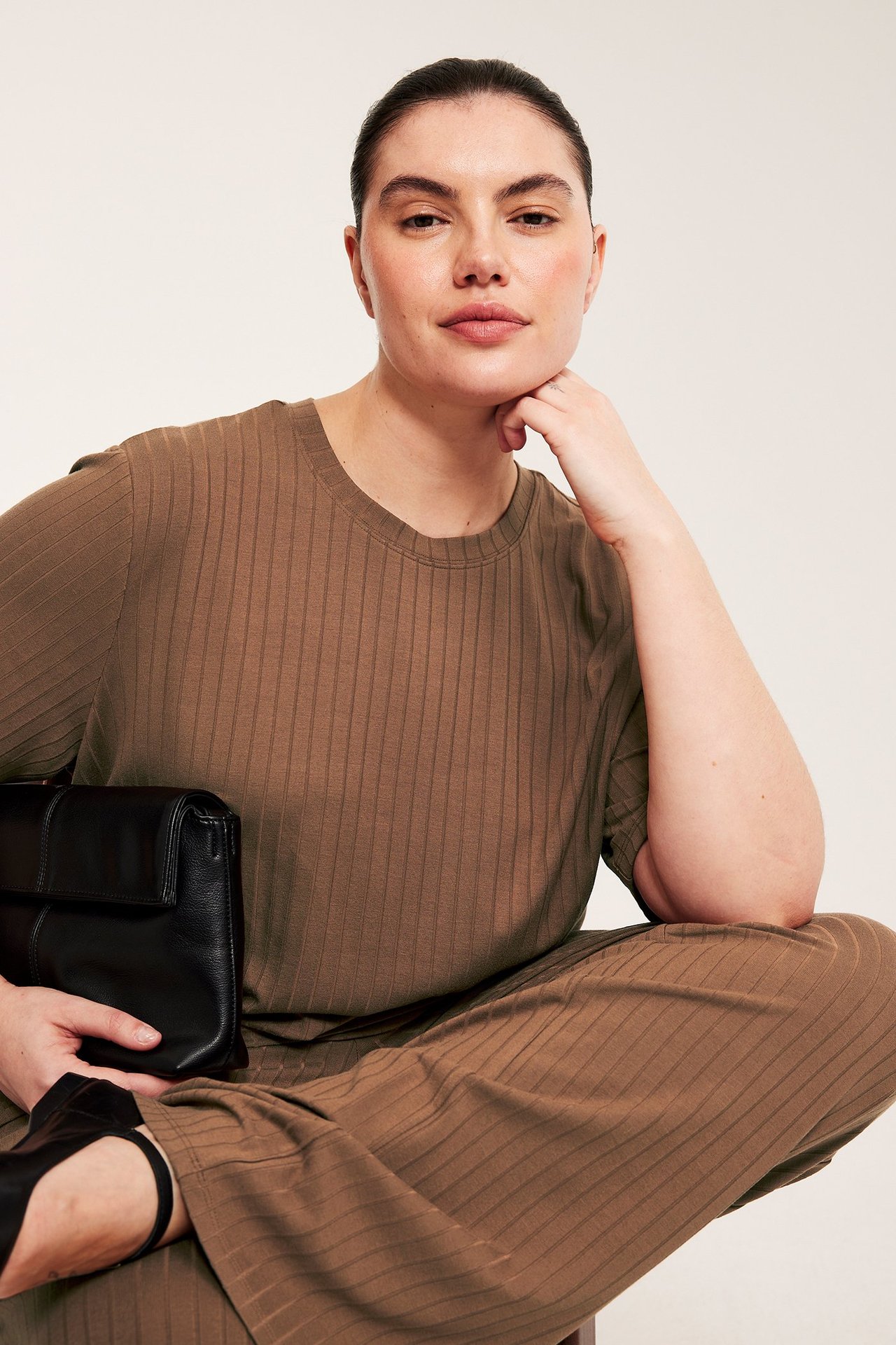Framifrån, kvinna i lång brun ribbad t-shirt, matchande byxor och håller en svart kuvertväska.