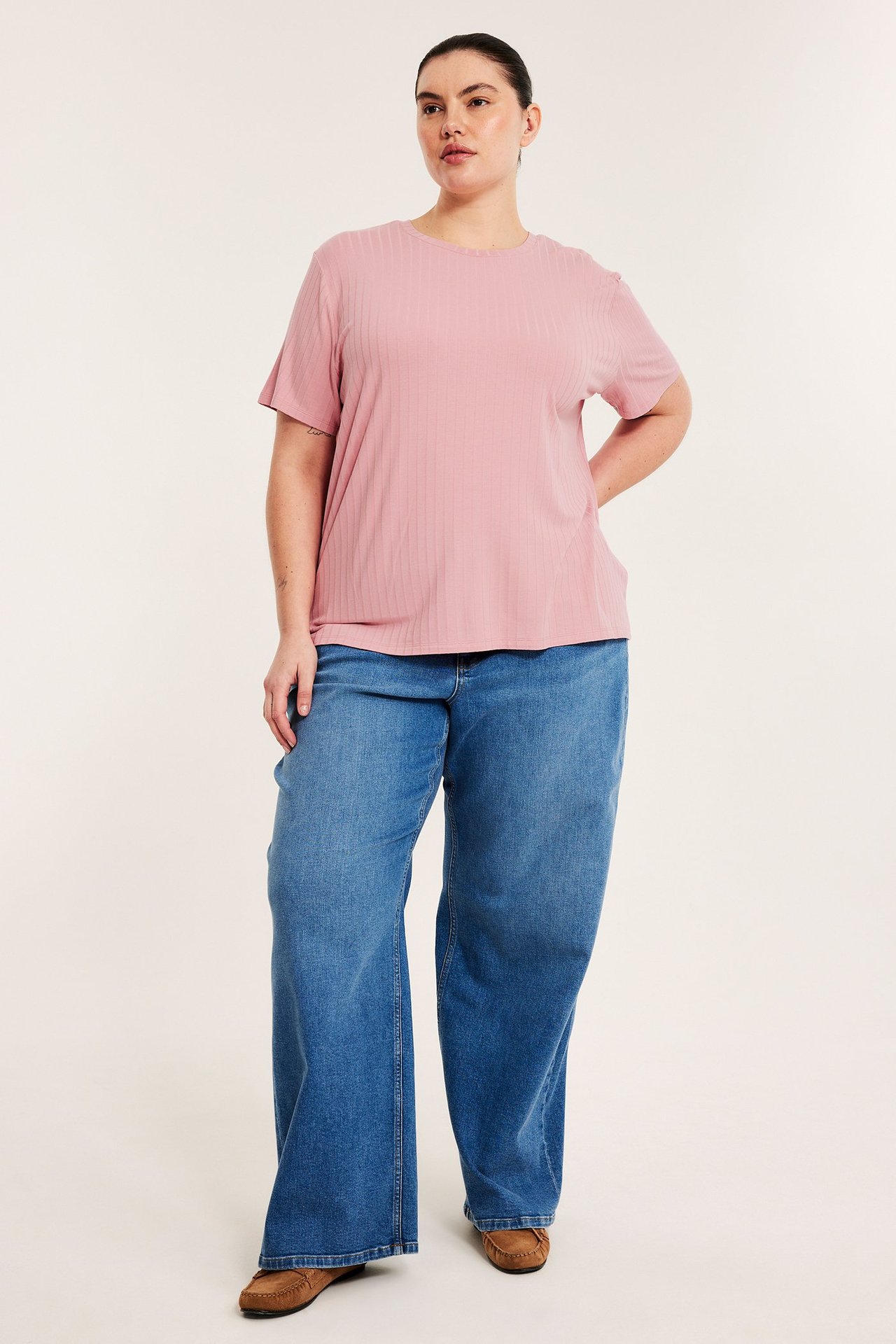 Nainen edestäpäin, yllään vaaleanpunainen ribbattu t-paita, siniset leveät farkut ja ruskeat mokkasiinit.