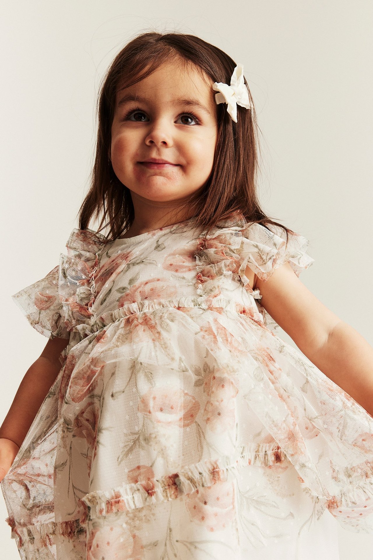 Edestä: Lapsi yllään luonnonvalkoinen kukkakuvioinen verkkomekko röyhelöhihoilla, hienovaraisin pilkuin ja valkoinen rusetti.
