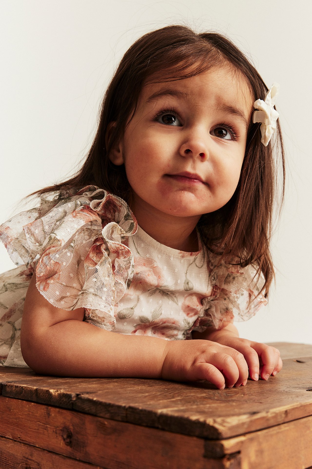 Lähikuva lapsesta luonnonvalkoisessa kukkakuosisessa verkkomekossa röyhelöhihoilla ja kukkahiussoljella.