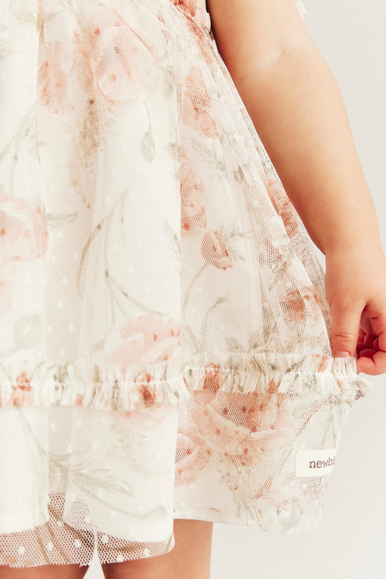 Lähikuva vauvasta offwhite-verkkomekossa, jossa vaaleanpunainen kukka- ja pistekuvio, pitäen röyhelöhelmaa.